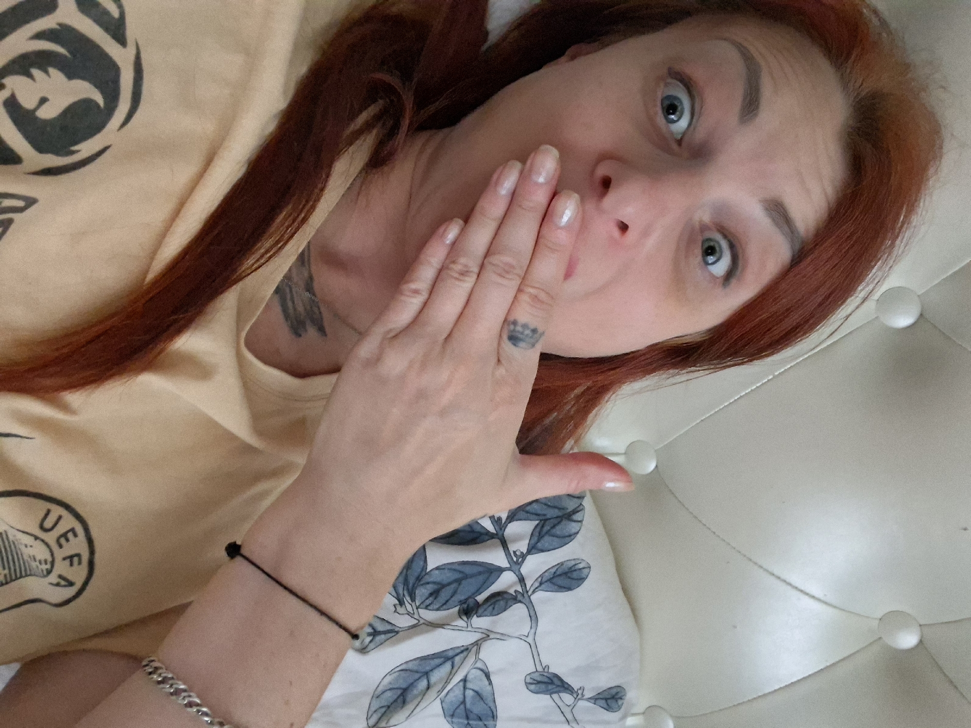 The image shows a woman with red hair lying in a bed. She is covering her mouth with her hand and has a surprised expression on her face. The bed has a tufted headboard and is covered with a pillow that has a botanical print. The woman is wearing a light-colored t-shirt with a graphic design. She is also wearing a thin black bracelet on her right wrist and a silver bracelet on her left wrist. A tattoo is visible on her left ring finger. There is another tattoo on her neck. The overall scene appears to be indoors, in a bedroom. There is no discernible information within the image that would allow for the determination of its location within the city of Myadzyel, Belarus.