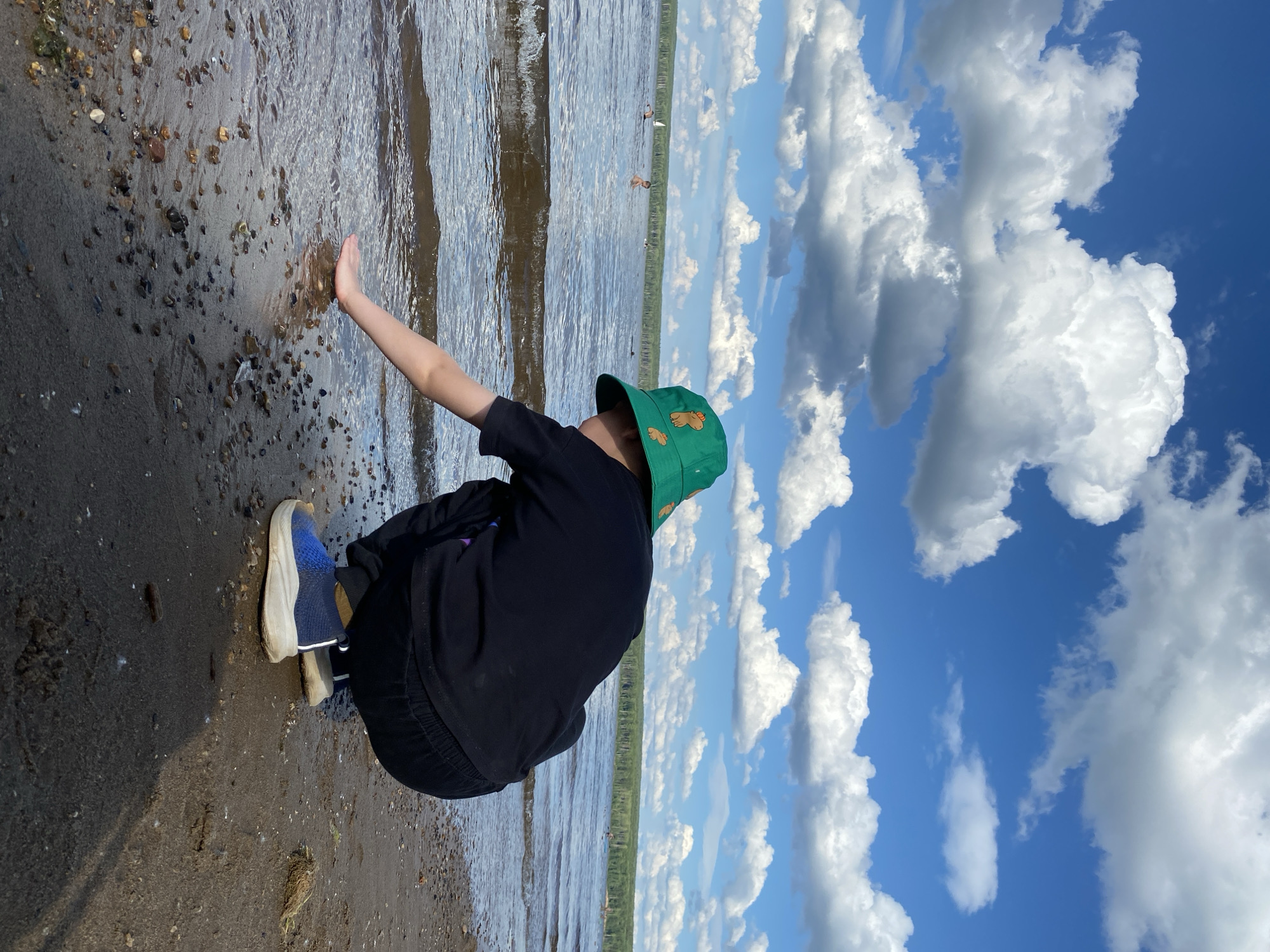 The image depicts a person, likely a child, crouching on the sandy shore of a body of water, possibly a reservoir or lake, with a tree line visible in the distance. The person is wearing a black t-shirt, black pants, and blue and white sneakers, along with a green bucket hat adorned with pineapple patterns. Their right hand is extended towards the water's edge, touching the ripples. Several other individuals are visible in the water, some appearing to be swimming or sailing small boats. The sky is a vibrant blue with scattered white, cumulus clouds. The environment suggests a natural setting, likely a recreational area near Chaikovsky, Russia, possibly along the Kama River, given the geographical context. The lighting indicates it is daytime, with sunlight casting a shadow on the sand.