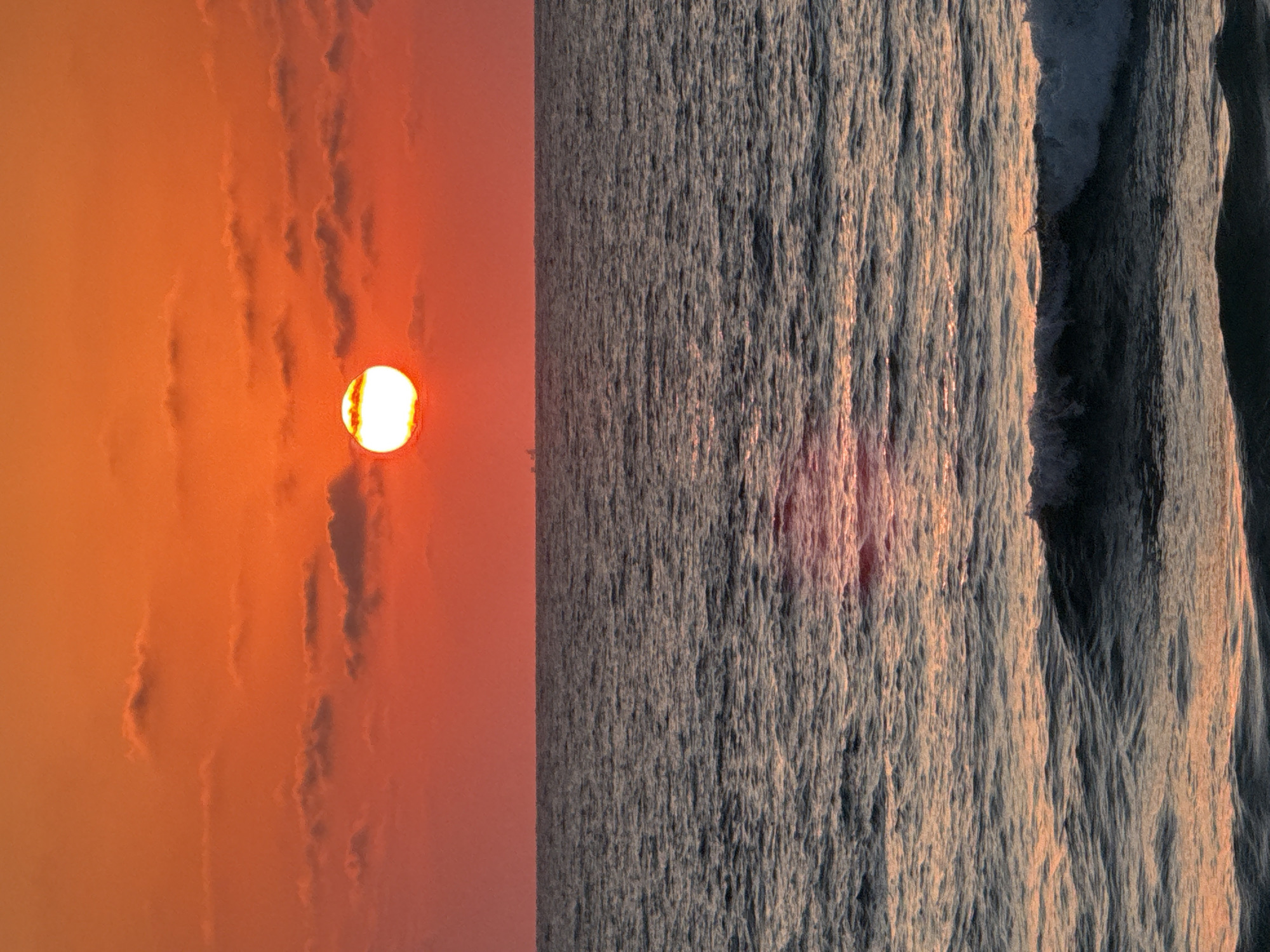 This media file shows a split view of a sunset over the ocean. On the left side, the sun is partially obscured by wispy clouds against a vibrant orange and pink sky. The sun itself is a bright, glowing orb with a yellow center. The right side of the image depicts the choppy surface of the sea, with waves reflecting the warm hues of the sunset. The water appears textured, with light and shadow creating a dynamic pattern. The overall impression is one of a serene and beautiful natural event. The time of day is clearly sunset, given the colors of the sky and the position of the sun. While the specific location is given as Tel-Aviv, Israel, there are no explicit visual cues within the image to confirm this. No people, objects, or text are visible in the image. The scene evokes a sense of peace and natural beauty.