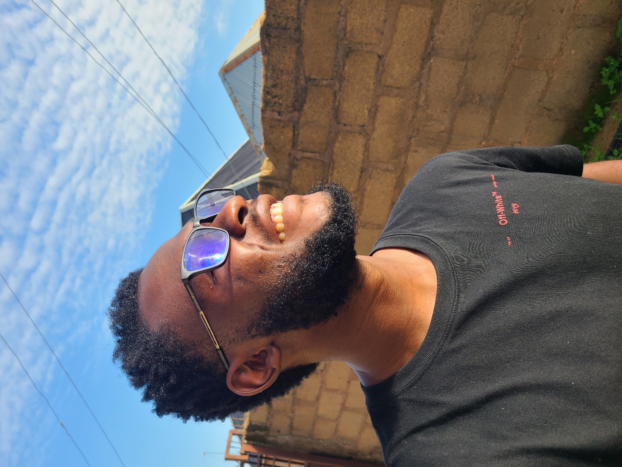 A medium shot profile of a Black man wearing black-rimmed glasses and a black t-shirt stands outdoors. He has a beard and is smiling, looking to his right. Behind him, a light brown brick wall runs horizontally across the frame. The sky above is blue with scattered white clouds. The scene is likely located in Kuru Karama, Nigeria.