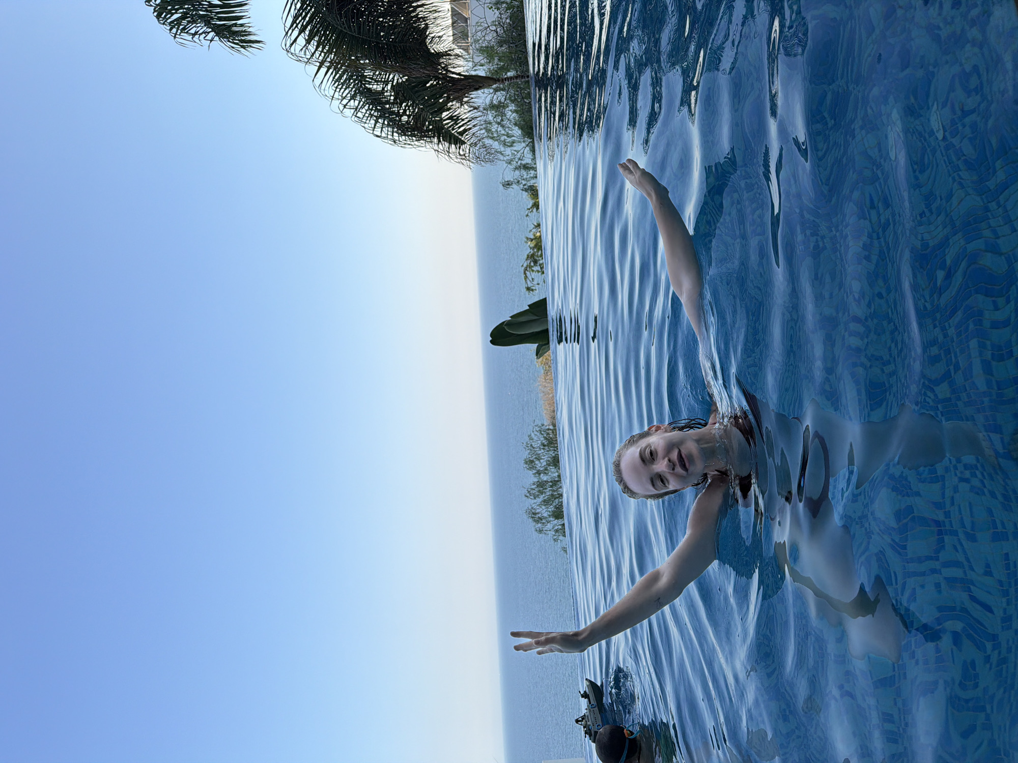In Agios Tychona Community, Cyprus, a woman with blonde hair is swimming in an infinity pool overlooking the sea. She is raising her left hand, making a "peace sign" with her fingers. The water is clear blue with a tiled bottom. In the background, the Mediterranean Sea stretches to the horizon under a clear blue sky. Palm trees and lush green foliage are visible on the hillside bordering the pool. To the left of the woman, another swimmer, wearing goggles and a snorkel, is partially submerged. A remote-controlled boat is also in the water near the second swimmer. The overall scene depicts a tranquil vacation setting.
