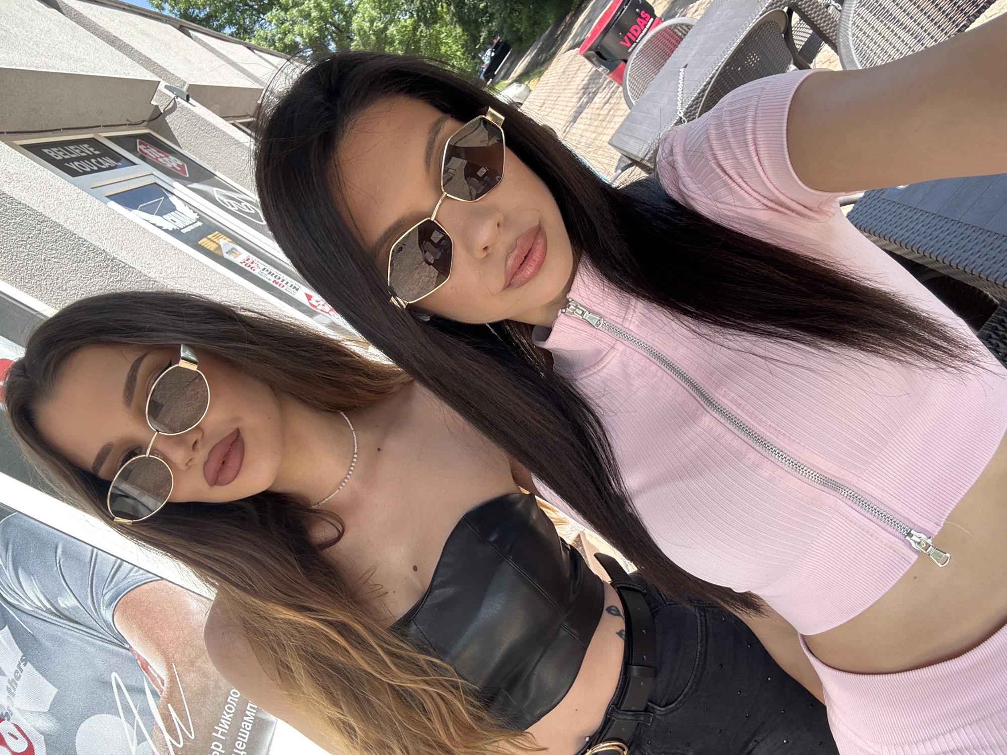 This is an outdoor, eye-level selfie featuring two young women on a bright, sunny day, likely in Sofia, Bulgaria.
The woman closer to the camera on the right has long, straight dark hair and is wearing a light pink, ribbed crop top with a silver zipper down the front. She pairs this with black bottoms and a dark belt. Her face is turned slightly to the left, and she has full, made-up lips. She wears gold-framed, hexagonal sunglasses with dark lenses, and her expression is posed and confident.
To her left and slightly behind, the second woman has long, wavy light brown/auburn hair and is wearing a strapless black crop top that appears to be made of a leather-like material. She also wears gold-framed, round sunglasses with dark lenses and a delicate silver chain necklace around her neck. Her lips are made up, and her expression is neutral.
In the background, there's a building with a textured grey facade. Several signs are visible on its windows; one prominently states "BELIEVE YOU CAN." Another partially visible sign shows "PROTEIN" with numerical information. Further back, outdoor tables and chairs made of a dark wicker-like material are present, along with a red cooler or container displaying the word "VIDAS." Part of a large poster with Cyrillic text, including "Браthers" (Brothers), "За Никола" (For Nikola), and "Шамп" (Champ), is visible next to the left woman. Green tree leaves indicate an outdoor setting, suggesting a cafe or street-side location. The overall atmosphere is casual and stylish.