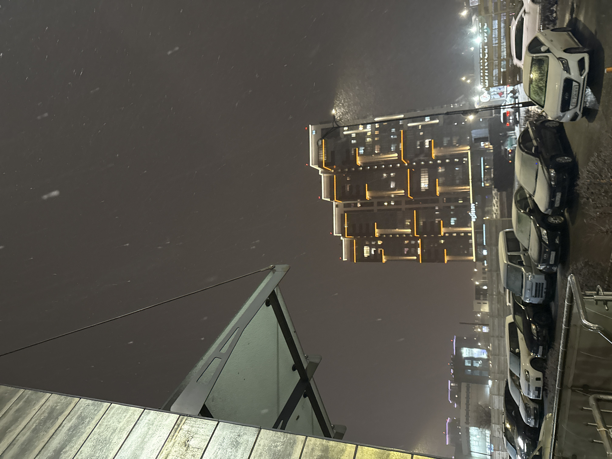 The photograph shows a snowy nighttime scene in Krasnoyarsk, Russia.
**Environment:**
* Heavy snowfall is occurring.
* Nighttime, illuminated by streetlights and building lights.
**Objects:**
* Multi-story building with illuminated windows and architectural lighting.
* Several parked cars, mostly white and dark colors.
* Street lights along the roadway.
* A section of a building or structure in the foreground, possibly a rooftop or balcony.
**Location:**
* Likely an urban area in Krasnoyarsk, Russia.