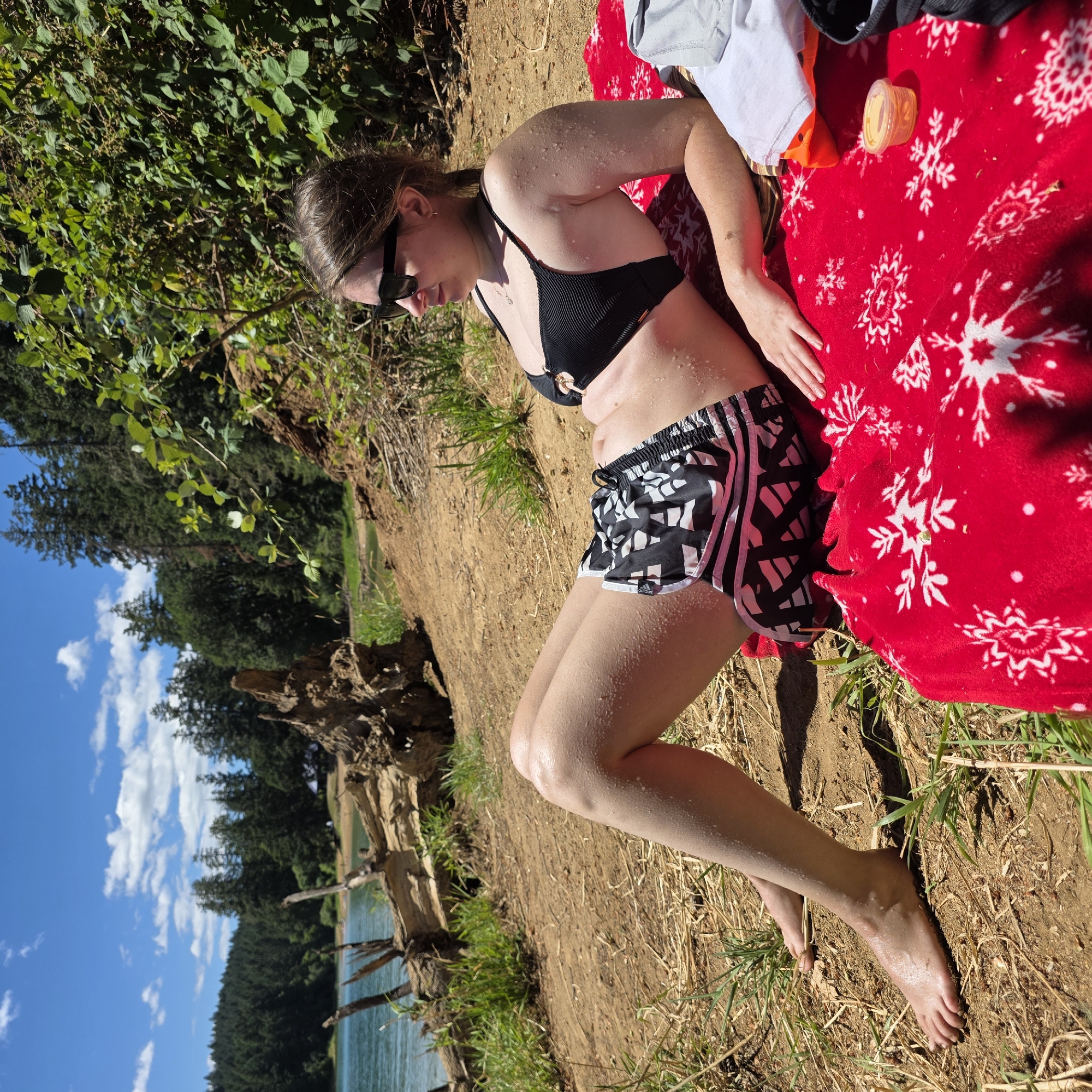 A young woman is lying on a red blanket with a snowflake pattern on a sandy bank next to a body of water. She is wearing a black bikini top and black and white patterned athletic shorts. Water droplets are visible on her skin, suggesting she has recently been in the water. She is wearing sunglasses and looking down. In the background, there are trees, a body of water, and a blue sky with white clouds. A piece of driftwood is visible on the left side of the image. A small orange plastic container is on the blanket. The image appears to be taken on a sunny day, likely in the afternoon, given the strong shadows. The presence of trees and a lake suggests an outdoor, natural setting. There is visible text on the shorts that reads "adidas".