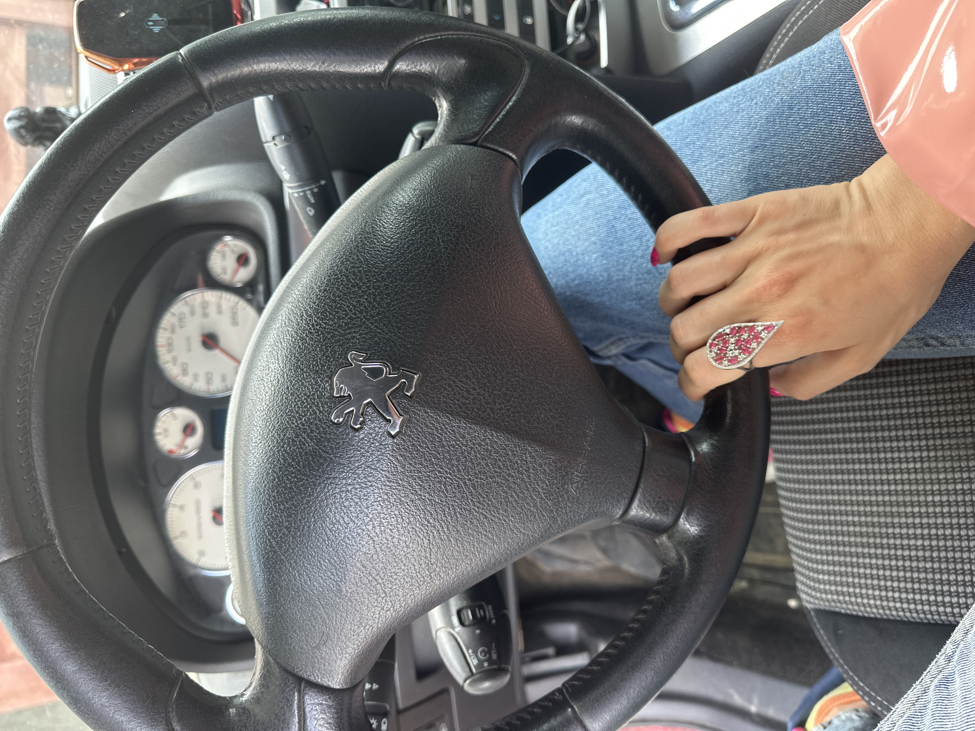 A close-up interior shot shows a black steering wheel with the Peugeot logo at its center. Behind the steering wheel, the dashboard gauges are partially visible. A woman's hand, adorned with a large ring featuring multiple small red stones in a teardrop design, is resting on the steering wheel. She is wearing a blue denim jacket and jeans. The seat cover is gray with a checkered pattern. The image does not indicate the city of Vladimir, Russia.