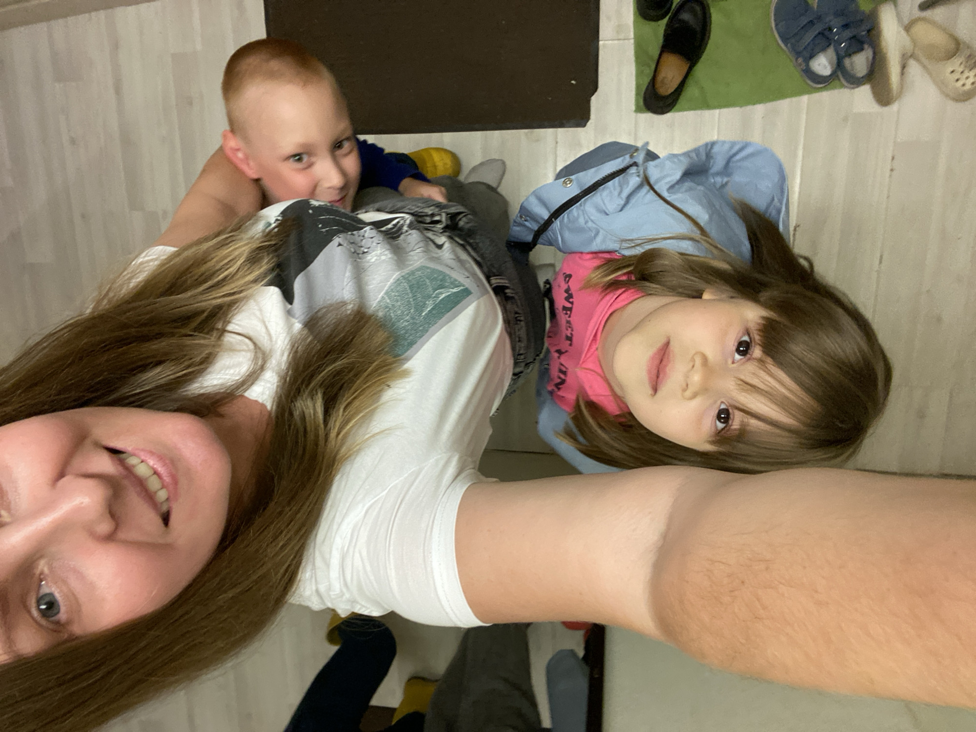 A selfie features a woman with shoulder-length brown hair, fair skin, and a visible smile, along with two children, a boy and a girl. The boy has a short, light-colored haircut and is wearing a blue shirt. The girl has brown hair with bangs and is wearing a pink shirt and a light blue jacket. They are positioned in front of a backdrop consisting of a tiled floor, a dark-colored mat, and several pairs of shoes, including slippers and black shoes. The location is identifiable as being in Noviy, Russia, although specific street details are not discernible from the image.