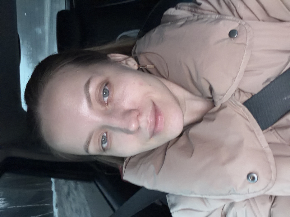 The image displays a female individual seated inside a vehicle, looking directly at the camera with a slight smile. She has light-colored eyes and light brown hair pulled back. Her face shows minimal makeup and faint blemishes. She wears a light tan or beige puffer jacket with a prominent collar, secured by a dark seatbelt across her chest and shoulder. A single gold-colored, rectangular drop earring is visible on her right ear. The vehicle's interior features dark elements, including a headrest visible behind her. Through the window on the left, a wintry outdoor scene is discernible, showing bare trees and snow-covered ground, suggesting a cold climate under dim, possibly overcast or twilight conditions. The specified location of Selevino, Russia, is not independently verifiable from the visual information within the image.