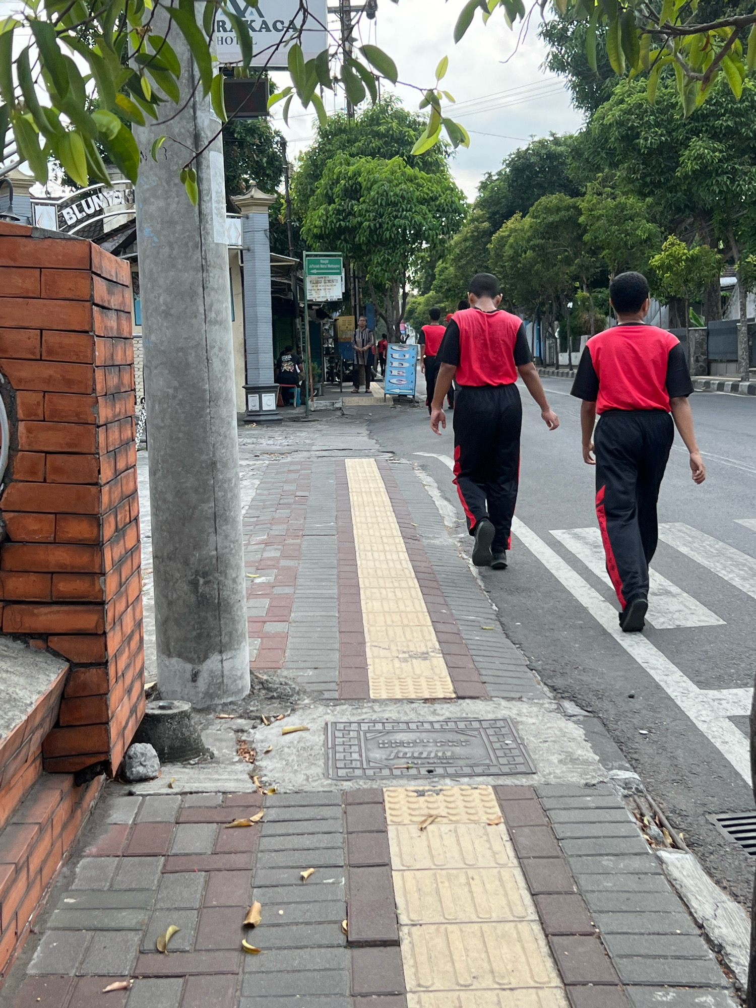 Two young males wearing matching red vests over black shirts and black pants with red accents are walking on a paved sidewalk in Yogyakarta, Indonesia. They are walking away from the camera, crossing a pedestrian zebra crossing on a street. The sidewalk is constructed with a pattern of colored paving stones, including textured yellow tiles, possibly for the visually impaired. A grey utility pole is situated on the sidewalk to the left of the frame, with a brick wall to its left. In the background, there are trees, buildings with signage, and other pedestrians. The sky appears overcast.