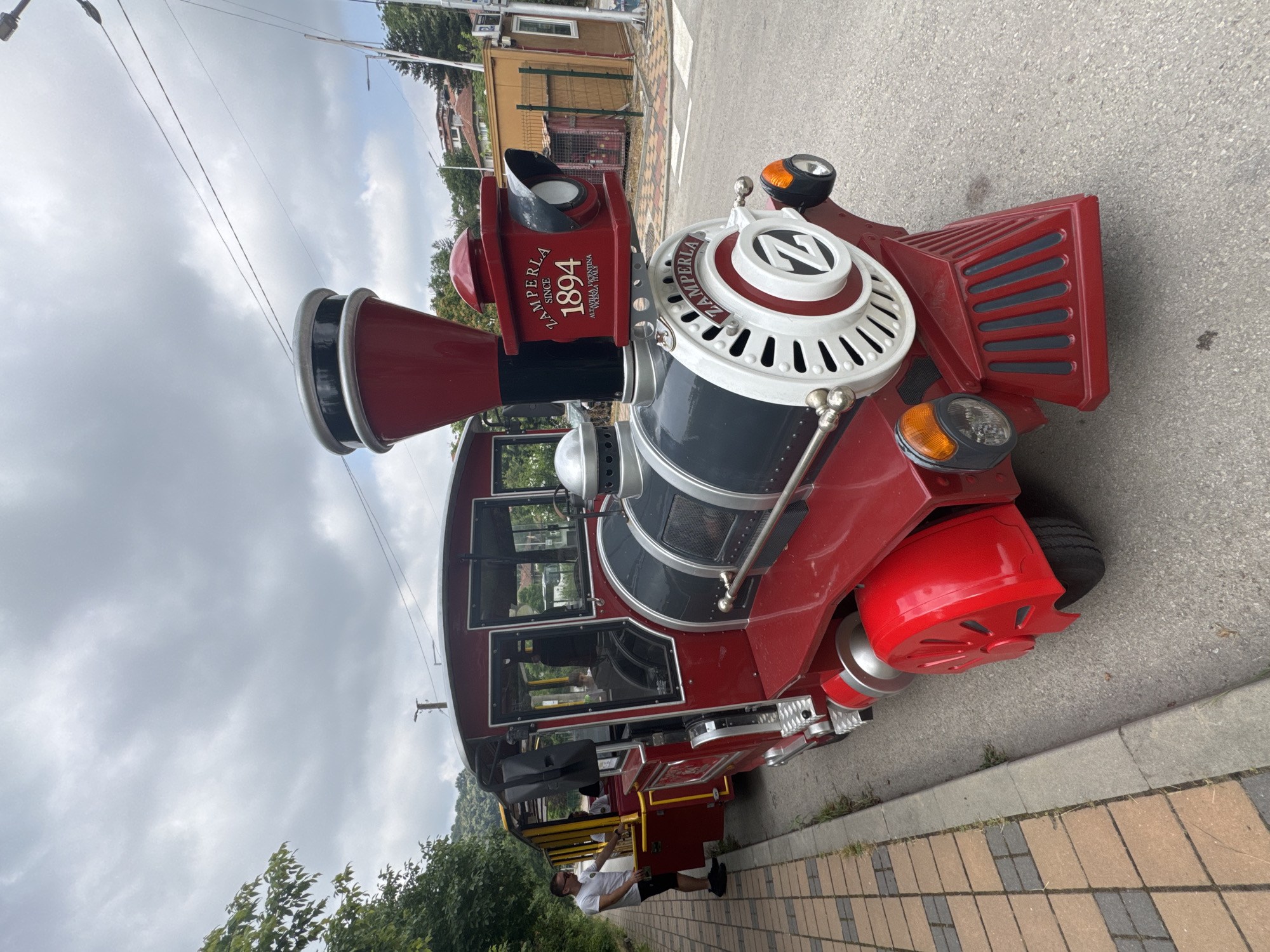 The image depicts a red and black miniature train, manufactured by Zamperla, parked on a paved surface next to a sidewalk in Veliko Tarnovo, Bulgaria. The train appears to be a tourist attraction, possibly a ride. A man in a white t-shirt and black shorts stands on the sidewalk, near the open door of one of the train's carriages, appearing to be an attendant or a passenger. The train has a decorative front with the Zamperla logo and "SINCE 1894" visible. The background includes trees, buildings, and a cloudy sky, suggesting an outdoor, urban setting. The overall scene conveys a relaxed, daytime atmosphere.