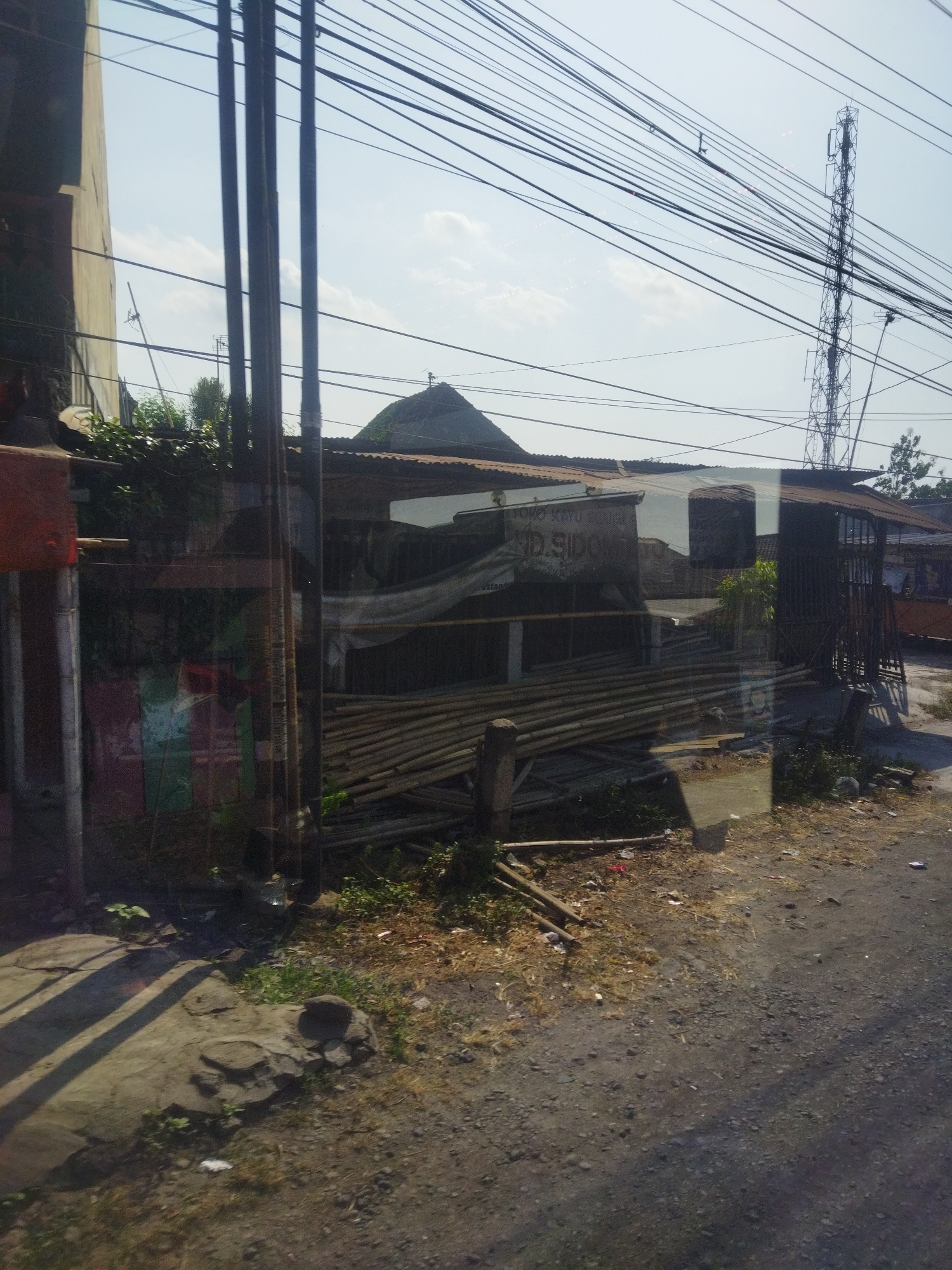 The image depicts a roadside scene in Nguter, Indonesia, likely a commercial or storage area. Dominating the upper portion of the frame is a dense network of overhead electrical wires crisscrossing against a pale blue sky with scattered clouds. To the right, a tall, metallic telecommunications tower stands prominently.
In the mid-ground, a low-lying structure with a corrugated metal roof appears to be a workshop or shop, partially obscured by debris and tarps. Stacked bundles of bamboo are visible in front of this structure, suggesting construction materials or a craft. A sign with some text, partially illegible due to reflections and distance, is attached to the building. To the left, a building with a lighter colored facade and visible vertical elements suggests a more permanent structure.
The ground in the foreground is a mixture of packed dirt, gravel, and scattered vegetation, with some concrete paving stones on the left. Small pieces of litter are visible on the ground, indicating a less-maintained area. The overall impression is of a functional, possibly informal, commercial or industrial environment along a road. No people are directly visible in the frame.