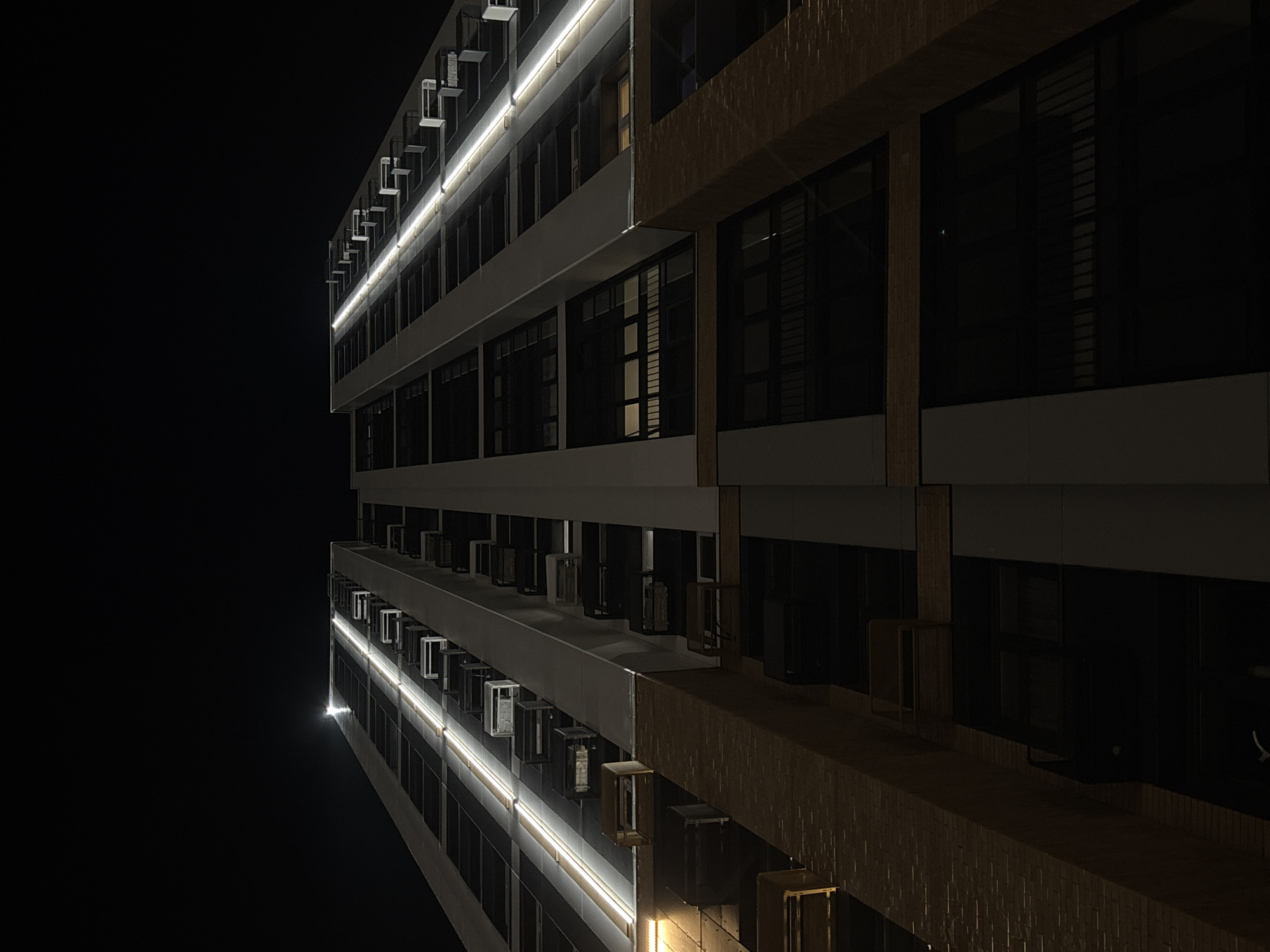 The image shows the corner of a multi-story building at night. The building appears modern, with a facade featuring rows of dark windows. Horizontal lights run along the edge of each floor, illuminating the building's exterior. There are several air conditioning units visible on the side of the building, between the windows. The surrounding environment is dark, suggesting a nighttime scene.
There is no information about the location within the city of Perm, Russia, nor any information about people, subjects, actions, or interactions.
