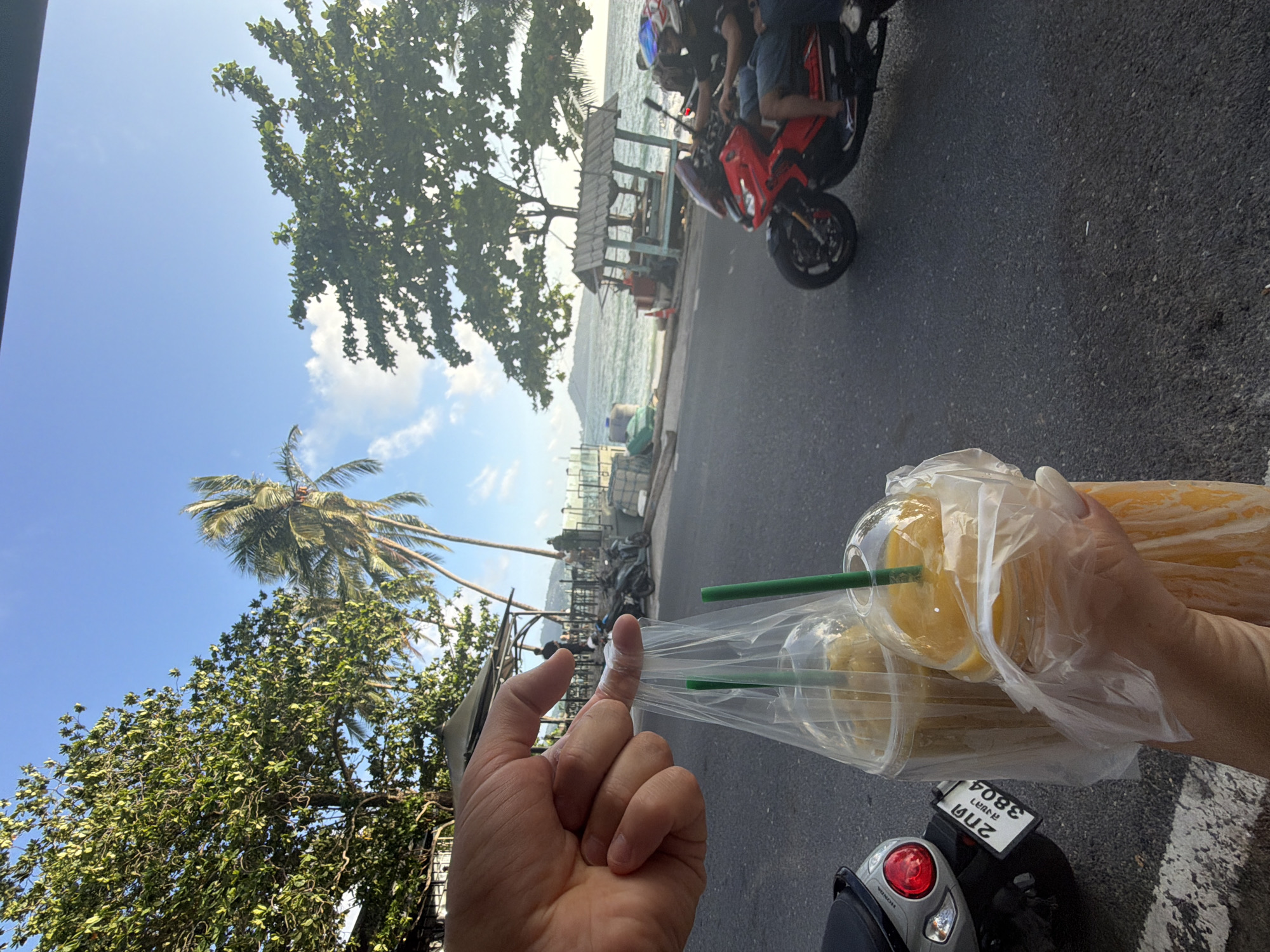 In Patong, Thailand, a close-up shot focuses on a hand holding two plastic cups filled with orange-colored beverages, each with a green straw, contained within a clear plastic bag. The cups are in the foreground, with the hand gripping the bag handle. Behind the drinks, a paved road is visible, bordered by a white line on the left. A silver scooter with a red taillight and a license plate reading "2 กล 3804" is parked on the left. Further in the background, a red motorcycle with two riders, one wearing a helmet, is in motion on the road. To the right of the road, a weathered wooden structure with a thatched roof stands near a body of water, possibly the ocean, with a distant island or landmass visible. Lush green trees, including palm trees, flank the scene under a bright blue sky with scattered clouds. Several people are standing or walking in the middle ground, near the wooden structure. The lighting suggests it is daytime, likely late afternoon due to the angle of the sun.