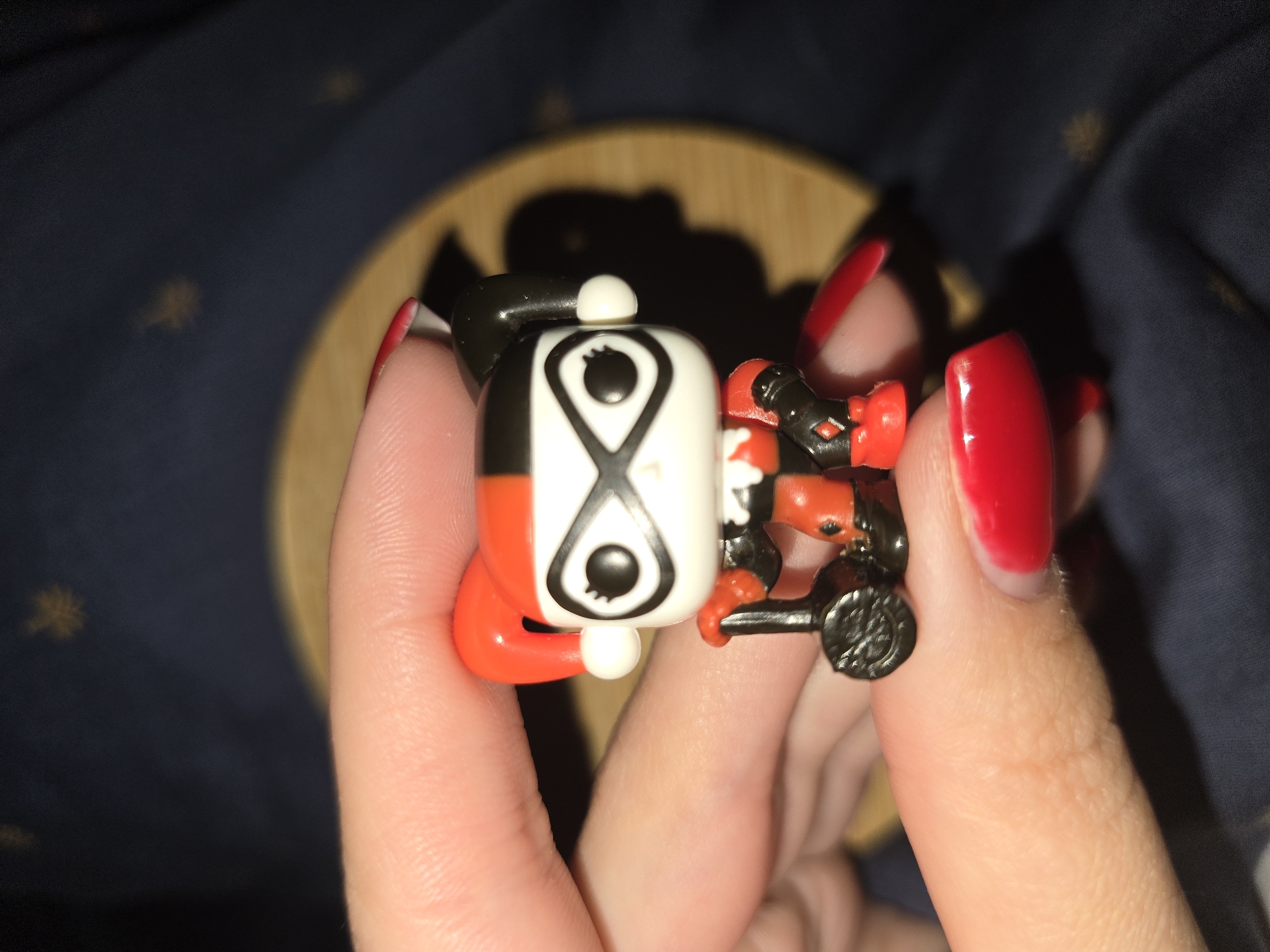 An extreme close-up shot shows a person's hand holding a small, stylized figurine. The figurine is of Harley Quinn, with her iconic red and black jester hat, mask, and costume. She is holding a black prop that appears to be a mallet. The hand has bright red fingernails. The background is dark and out of focus, with a circular wooden object visible. The scene appears to be indoors, as indicated by the lighting and shadows. There is no information available about the location being Petropavlivska Borshchahivka, Ukraine.