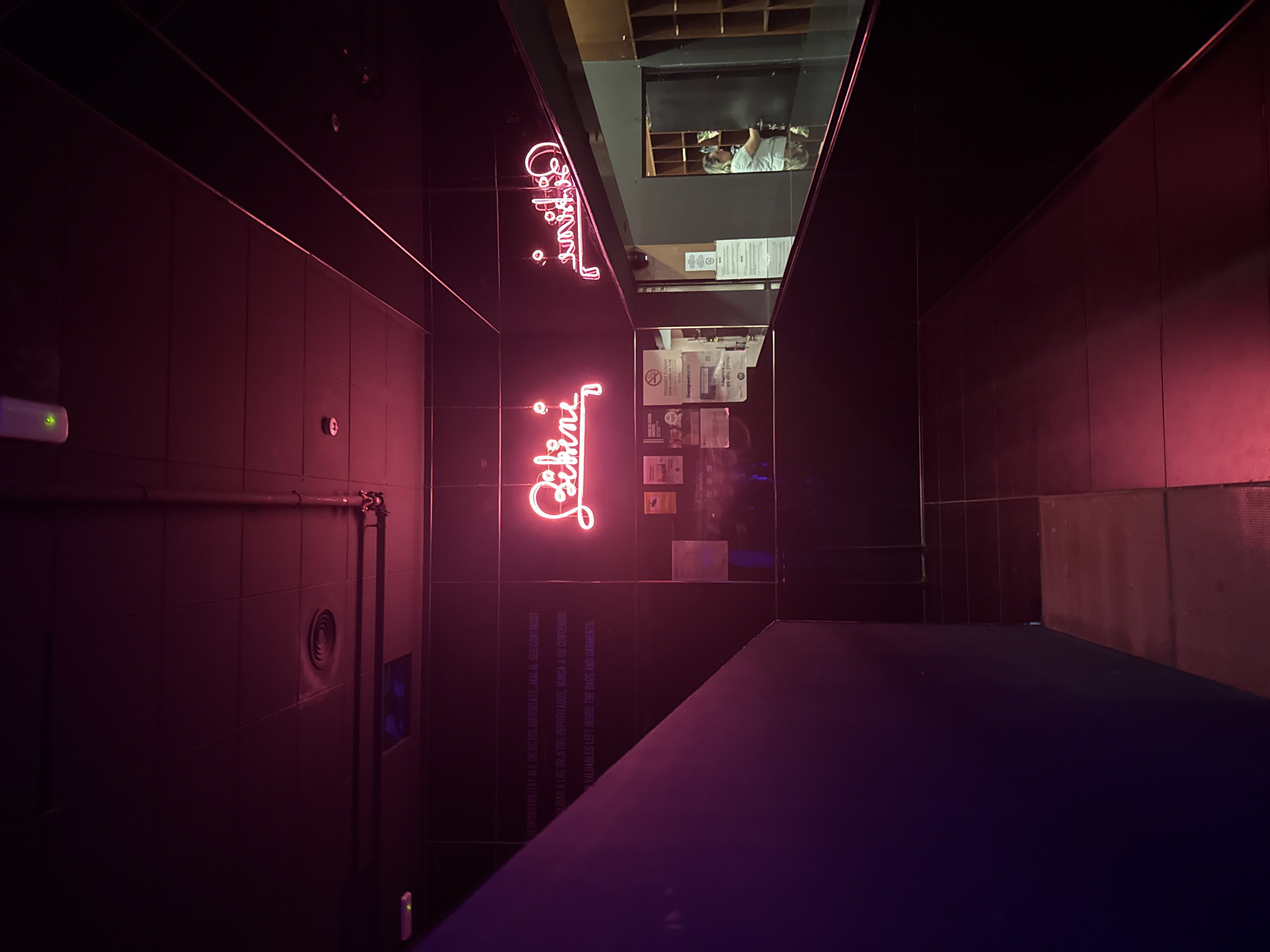 The image shows the interior of the "Bikini" nightclub in Barcelona, Spain. The walls are dark, tiled, and illuminated by a neon pink sign spelling out "Bikini." A row of informational posters are visible on the wall, along with pipes and an air vent. A blue-purple object is also visible, and a reflection of a person can be seen in a mirrored section above the neon sign.