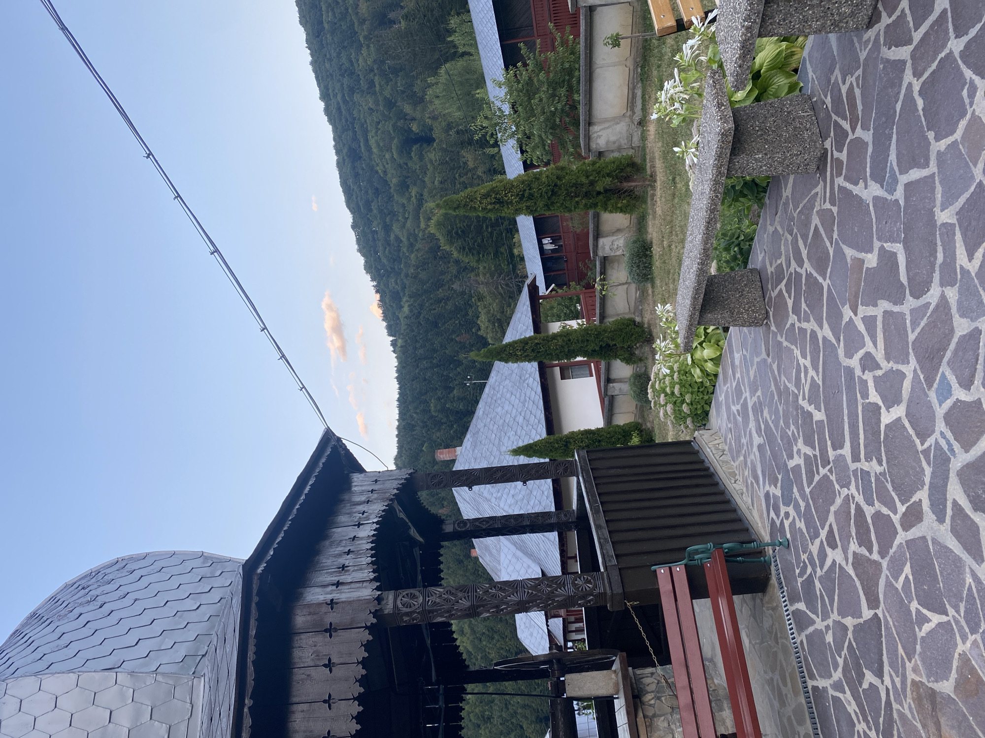The image depicts a serene outdoor scene with traditional architecture and natural surroundings, likely in Crăcăoani, Romania. In the foreground, a stone-paved patio features two concrete benches with a pebble-like texture. To the left, a red wooden bench is situated next to a dark wooden structure with intricate carvings, possibly part of a well or gazebo, topped with a grey, hexagonal-patterned roof. Behind these elements, several buildings with grey tiled roofs are visible, nestled amongst lush greenery. Tall, slender evergreen trees punctuate the landscape. In the background, a densely forested hill slopes upwards under a clear sky with a few wispy clouds. A power line stretches diagonally across the upper portion of the frame. The overall atmosphere is tranquil and rustic.