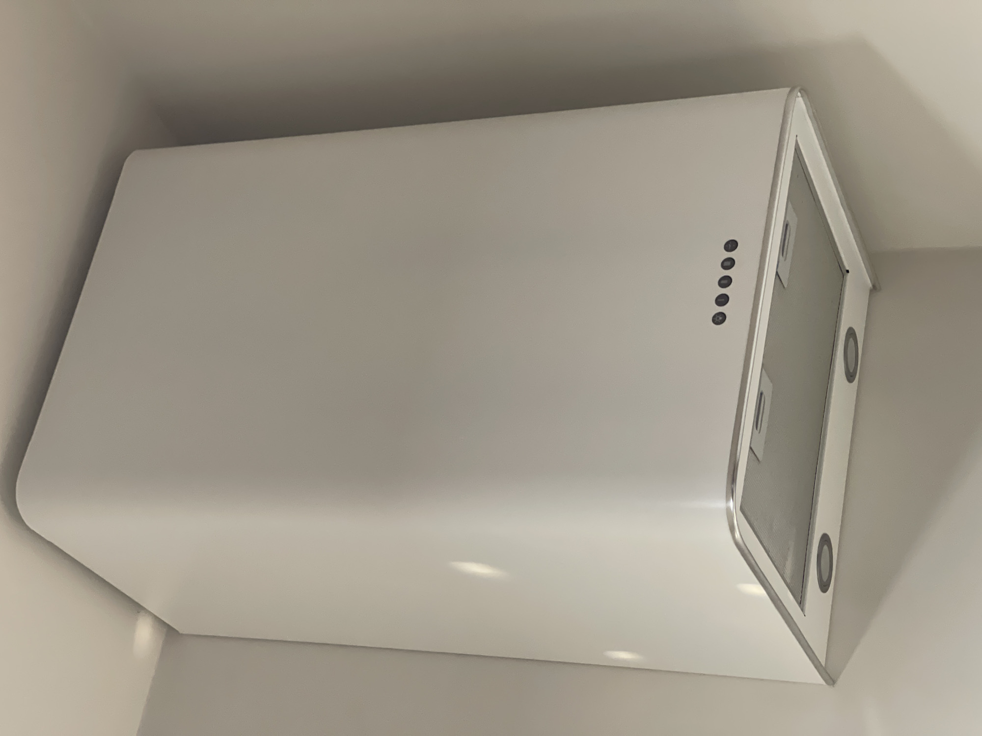 An angled, close-up shot captures a sleek, modern kitchen range hood in a white or off-white finish. The hood is mounted against a wall and extends into what appears to be a corner or against another wall, highlighted by subtle shadows. Its design features a rectangular body with gently rounded top edges, framed by a slim metallic trim along the front and side.
On the front-right panel of the hood, five circular black control buttons are arranged vertically. From top to bottom, the visible symbols suggest functions such as a light (light bulb icon), fan speed adjustment (likely up and down arrows or power levels), and a power on/off button.
The underside of the hood reveals two rectangular, metallic mesh filters, designed to capture grease and impurities. Integrated into the underside, and partially visible around the filters, are circular outlines that suggest built-in lighting fixtures. Reflections on the smooth surface of the hood indicate indoor lighting, possibly from recessed lights in the ceiling or external light sources. The surrounding walls are also a light, neutral color, complementing the hood's minimalist aesthetic. No people or visible text are present in the image.