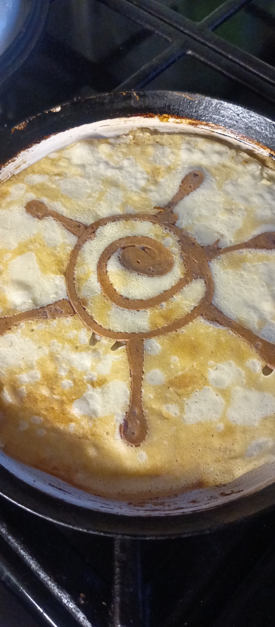 A circular pancake with a spiral design in a frying pan on a stovetop. The pancake is light yellow with brown accents, and the spiral design is brown. The frying pan is black with a white interior. The stovetop is black.