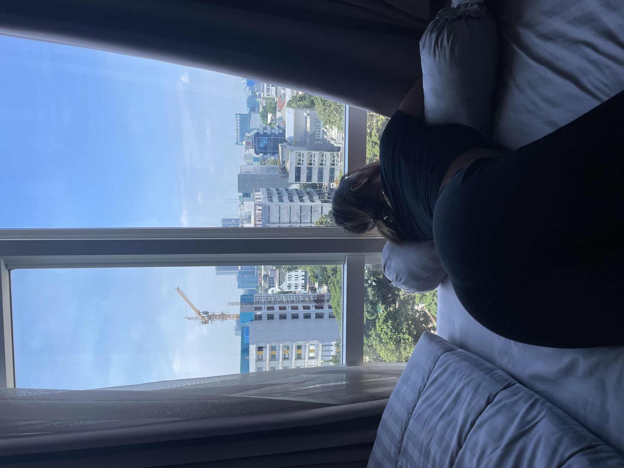 A person is lying on a bed near a window. Outside the window is a cityscape, indicating a dense urban environment. In the distance is a yellow construction crane. Based on the city architecture, the location appears to be in Central Jakarta, Indonesia. The person's hair is light brown. They are wearing a dark shirt and dark pants. Pillows are positioned around the person, and there is a striped comforter on the bed. Natural light streams into the room through the window.