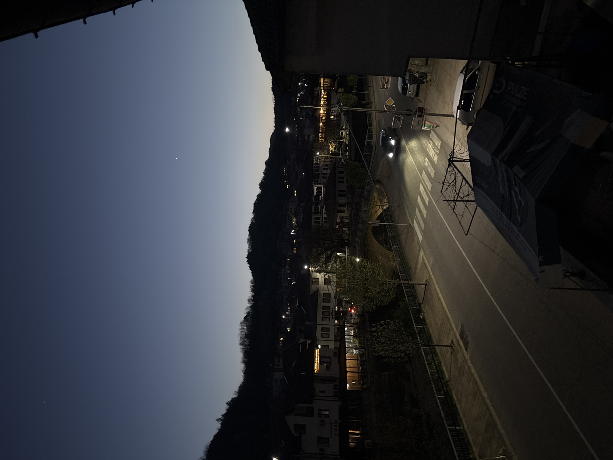 The image captures a dusk or early night scene in a town, likely Zlatograd, Bulgaria, viewed from an elevated perspective, possibly a balcony. The sky transitions from a dark blue-grey at the top to a lighter hue towards the horizon, with a single bright celestial body (likely a star or planet) visible. A dark, forested hillside, characteristic of mountainous terrain, forms the backdrop on the right.
In the foreground, a part of a dark structure, possibly a roof or overhang, frames the top. Below this, a street with a white dividing line runs diagonally, illuminated by streetlights. Two cars are visible on the road, both with headlights on, one appearing closer and moving towards the viewer. A pedestrian crossing is faintly discernible on the asphalt.
Along the street, various buildings, including multi-story structures, are visible, with many windows and facades illuminated, suggesting residential or commercial activity. Trees, some bare and some with foliage, are scattered among the buildings and alongside the road. A large billboard or sign structure stands prominently on the right, partially obscured by shadow. Traffic signs are also present near the road.
The time of day is clearly evening or early night, indicated by the dark sky, artificial lights, and the gradient of the sky reflecting fading daylight. The weather appears clear. The scene depicts the routine evening life of a small town nestled in a valley, with lights providing warmth against the encroaching darkness of the natural landscape.
Visible text on the prominent billboard includes "PULZE", possibly "Sevan" below it, and "Davidoff". On one of the buildings, the letters "TOHH" or a similar word can be seen.