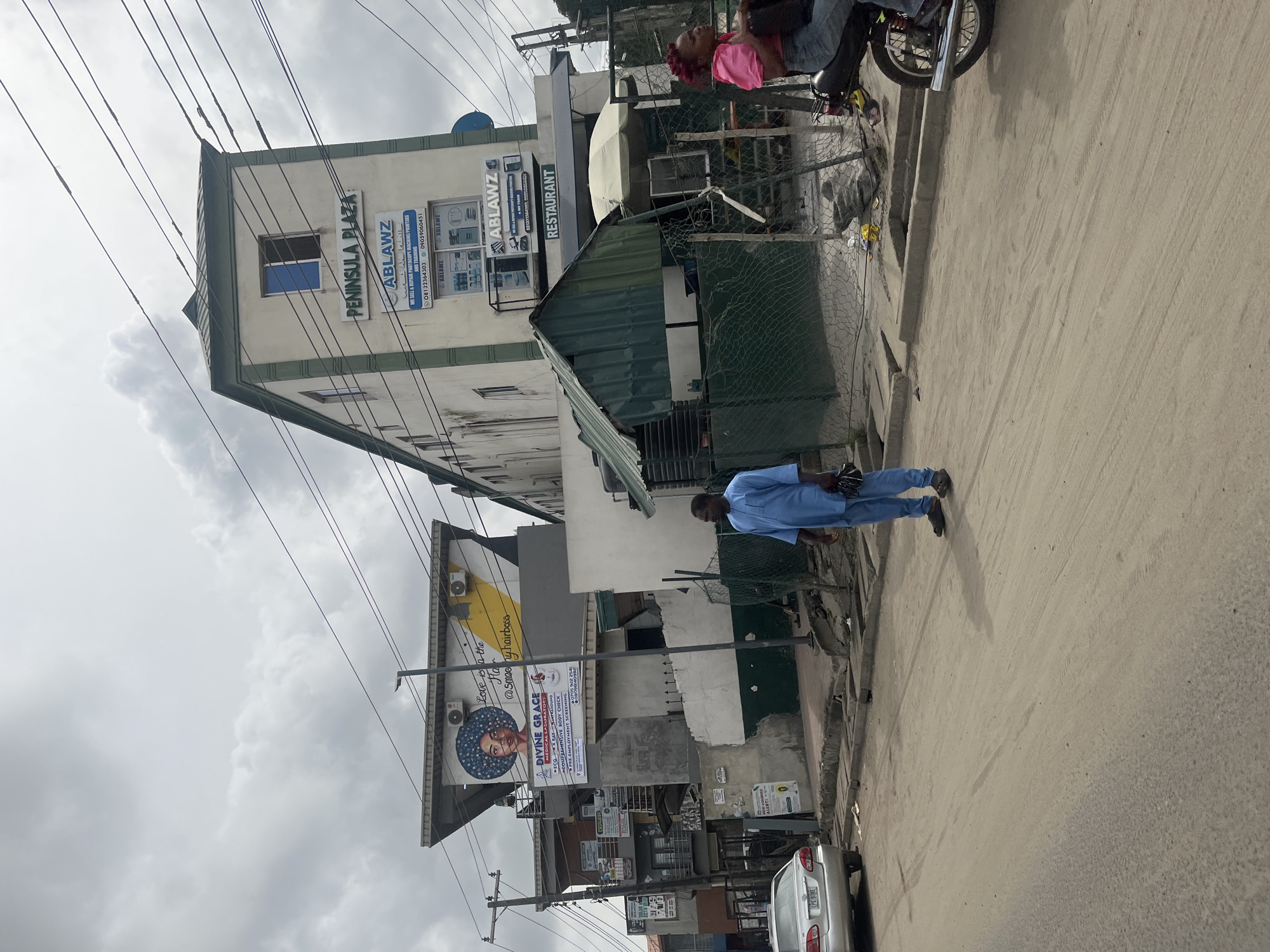 Here is a description of the image:
The image depicts a street scene in Lekki, Nigeria. On the left, a multi-story building bears the sign "PENINSULA PLAZA: ABLAWZ", advertising photocopy machine sales and repair services, and there's an advertising sign that reads "DIVINE GRACE MEDICAL LABORATORY." On the street, a man in a light blue outfit walks along the pavement. In the background, a motorcycle is parked with two people on it. The sky is overcast. A car is parked along the curb. Power lines crisscross above.