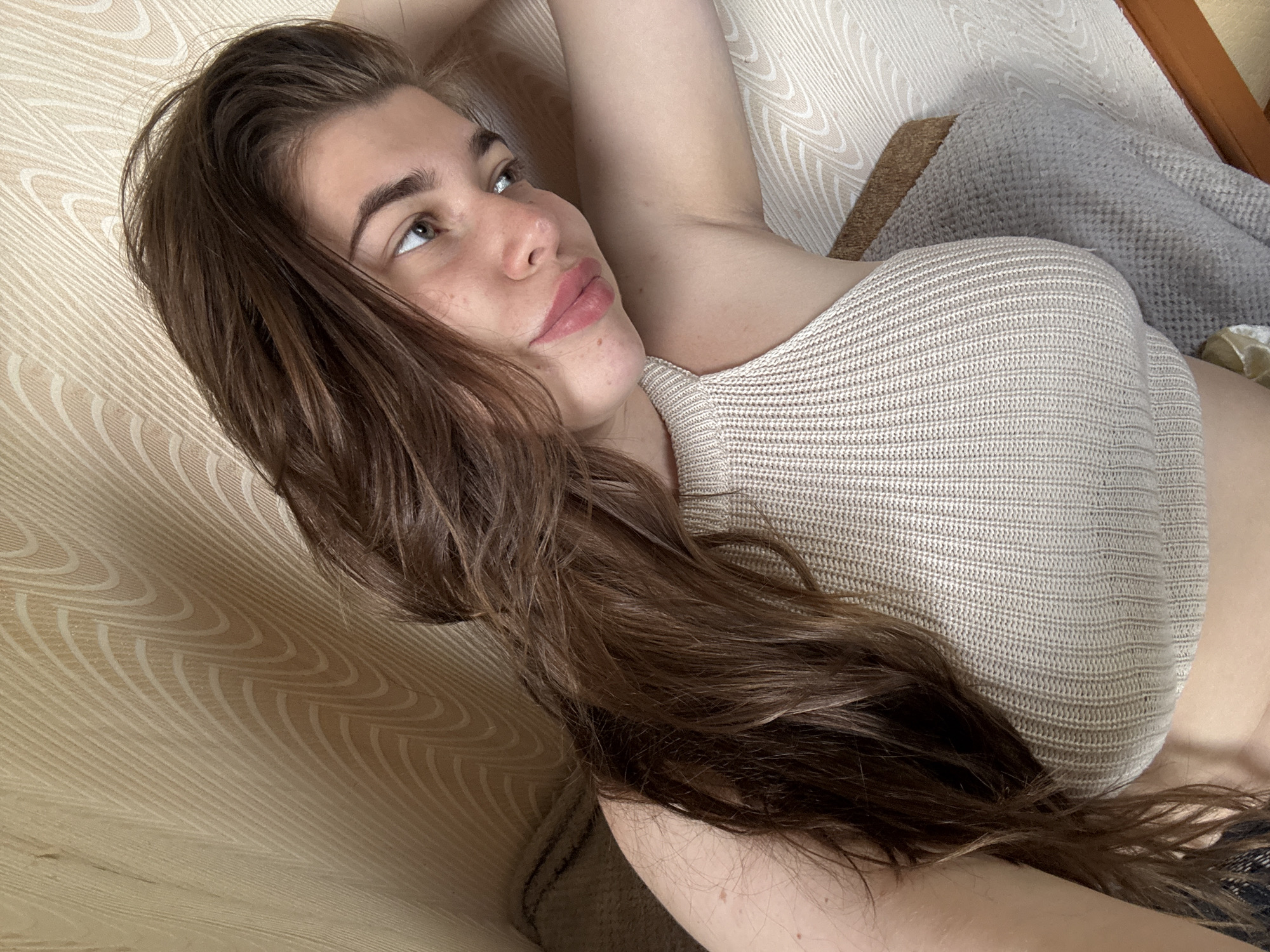 A young woman with long, dark brown hair is lying down, looking to her right. She is wearing a beige, ribbed, halter-neck top. Her right arm is bent, and her hand is resting on her head. She has light skin with some freckles. The background consists of a textured, beige wall with a wavy pattern, and a blurred grey blanket or sofa. The scene appears to be indoors. There is no indication of the city of Мещёры, Russia in the image.