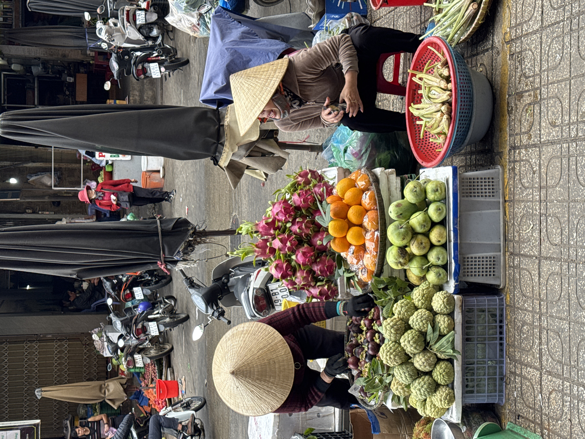 This is a full shot of an outdoor market stall in Da Lat, Vietnam. Two women are present, both wearing traditional conical hats. One woman, in the foreground left, is crouched down, wearing a dark-colored patterned shirt and black pants, and appears to be tending to a basket of fruit. She is also wearing black gloves. The other woman is sitting on a red stool to the right, wearing a beige hooded jacket and black pants. She is looking to the side and appears to be speaking.
The stall is laden with fresh produce. In the foreground, there are several baskets of various fruits, including what appear to be mangosteens, custard apples, dragon fruit, and oranges. There are also bundles of green vegetables, possibly okra and long beans, in the lower right corner. In the background, several motorcycles are parked, and there are large black umbrellas providing shade over the market stalls. The ground is paved with patterned tiles. The overall scene depicts a vibrant market atmosphere.