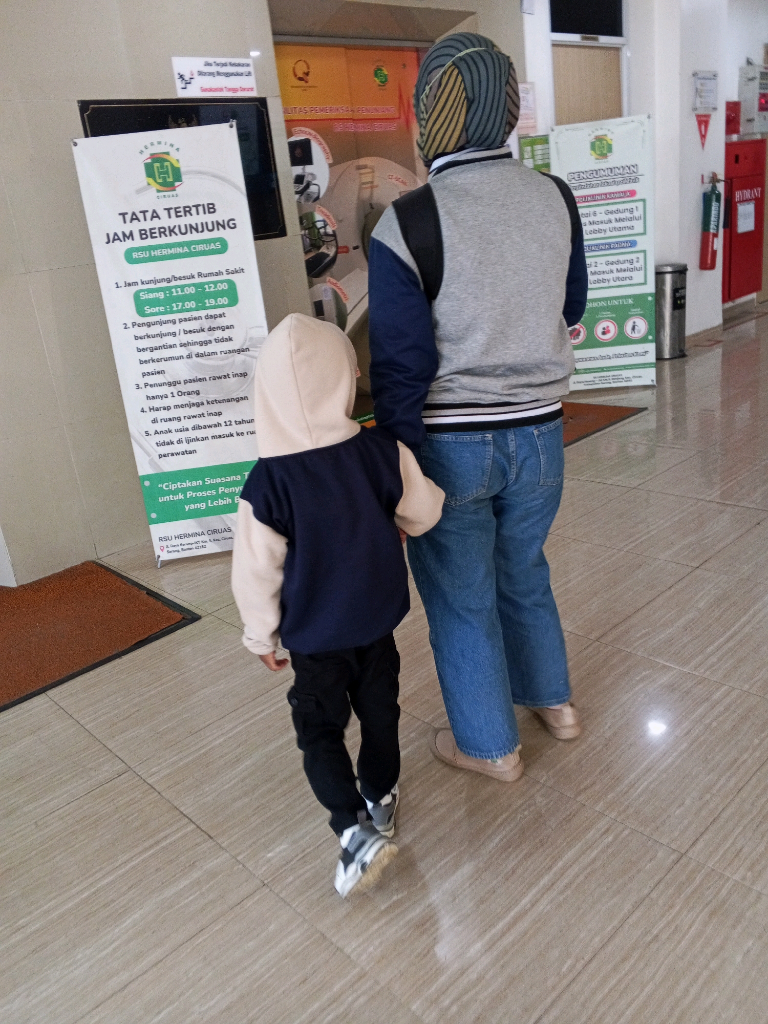 Two individuals, an adult and a child, are standing in a hallway. The adult, viewed from behind, wears a gray and blue jacket, blue jeans, and light-colored flat shoes. The child, also viewed from behind, wears a beige hooded jacket, black cargo pants, and black and white athletic shoes. They appear to be walking together.
Behind them are several informational posters. One poster, titled "TATA TERTIB JAM BERKUNJUNG," outlines visiting hours and rules for RSU Hermina Ciruas. Another poster, titled "PENGUMUMAN," provides information regarding clinic access.
The environment is a hospital hallway with tiled flooring. To the right of the posters, a red fire hydrant is visible mounted on the wall. The lighting suggests an indoor setting. The presence of the hospital name indicates the location is RSU Hermina Ciruas in Ciruas, Indonesia.