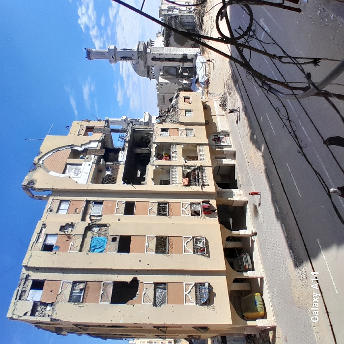 The image depicts a devastated urban street scene in Khan Yunis, Palestinian Territory, under a bright blue sky with scattered white clouds. The immediate foreground is partially obscured by dark wires and metal framework.
A multi-story, beige-colored apartment building dominates the left side of the frame. This building exhibits extensive structural damage, including numerous circular impact holes pockmarking its facade, large sections of walls and roofing completely destroyed or missing, and broken windows. Clothes are visibly hanging from several balconies, some with white railings. Underneath the building's ground-floor arches, a white sedan and a yellow tarp-covered object are parked.
A paved two-lane road runs horizontally across the mid-ground. On the sidewalk adjacent to the damaged apartment building, a small individual dressed in a red shirt is standing.
In the mid-ground to the right, a white mosque with a tall minaret and a dome is visible. The mosque also displays significant damage, with visible holes and missing sections in its structure and dome. An individual is seen walking through a debris-strestrewn area located between the road and the mosque.