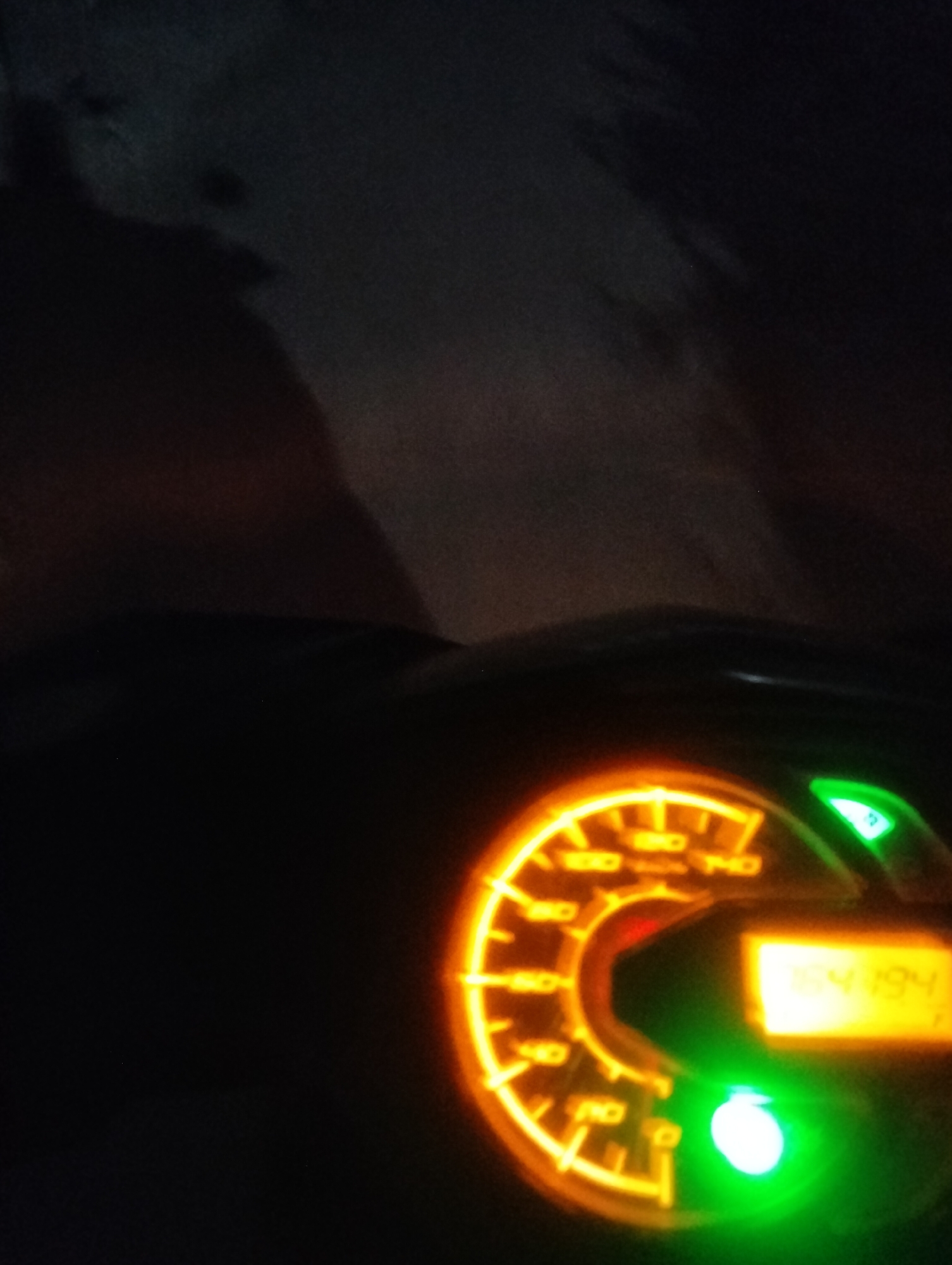The image shows a close-up of a motorcycle's digital instrument cluster at night. The cluster features an analog speedometer with orange backlighting, displaying markings up to 140 km/h. A digital display shows the number "164194". To the right of the speedometer, there is a green indicator light, possibly for turn signals, and a green illuminated button or light at the bottom. The background is dark and appears to be a blurry outdoor environment, suggesting the motorcycle is parked or moving in low-light conditions. The city of Lhokseumawe, Indonesia is not depicted in the image.
