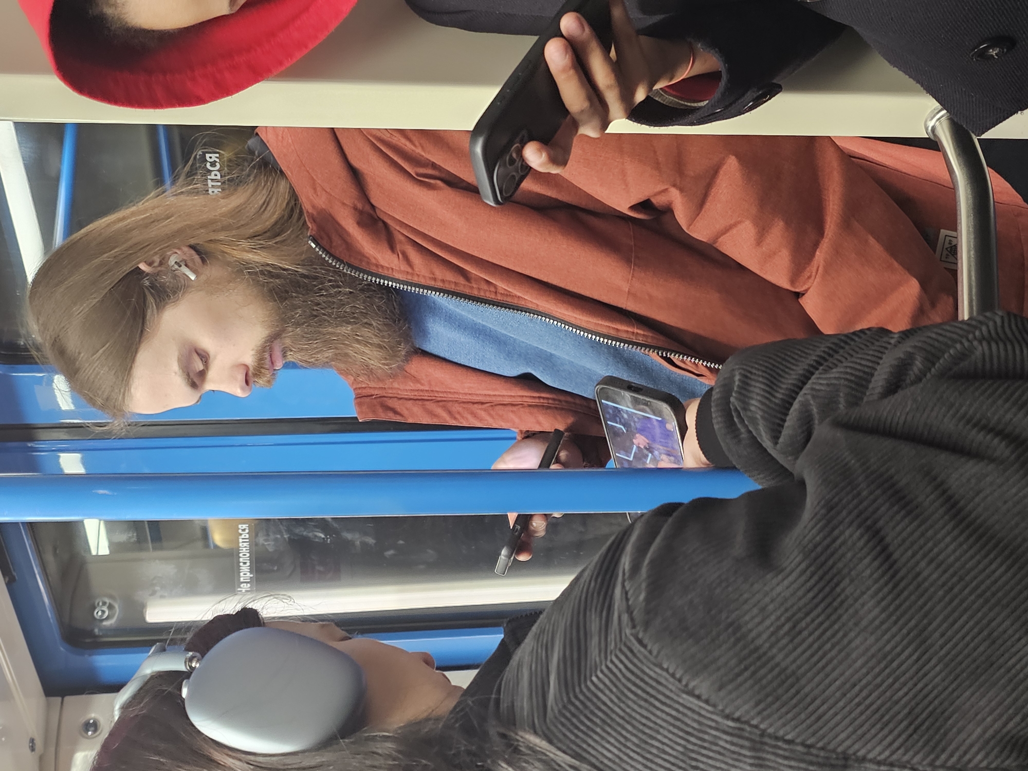This image captures an interior view of a public transport vehicle, likely a train or subway car, in Khimki, Russia. The scene is dominated by several passengers, with a man in the foreground at the center.
The man, with long light brown hair and a full beard, has his eyes closed or cast downwards. He wears a rust-colored zippered jacket over a blue top and has a white earbud in his left ear. To his right, partially obscured by the frame, another individual holds a dark-cased smartphone. Visible on the screen are colorful graphics, suggesting they might be watching a video or playing a game. Above the man's head, another person's hand holds a black smartphone with a triple-camera setup, indicating a newer model. Further to the top-left, only a hint of a red hood is visible.
In the bottom-right, a person with dark hair is seen wearing large, light grey over-ear headphones. They are dressed in a dark, ribbed corduroy-like jacket. Another smartphone is partially visible near this person's hand.
The setting features prominent blue handrails or bars running horizontally across the vehicle's interior. A light-colored panel forms the ceiling or upper wall, and windows with reflections are visible. The lighting appears bright and uniform, suggesting either daytime travel or a well-lit interior.
A notable detail is visible text on a window, which reads "НЕ ПРИКОСНАТЬСЯ" (NE PRIKOSNATSYA), meaning "DO NOT TOUCH" in Russian, a common safety or warning sign in public spaces. The overall activity suggests passengers commuting or traveling, engaging with their personal devices.