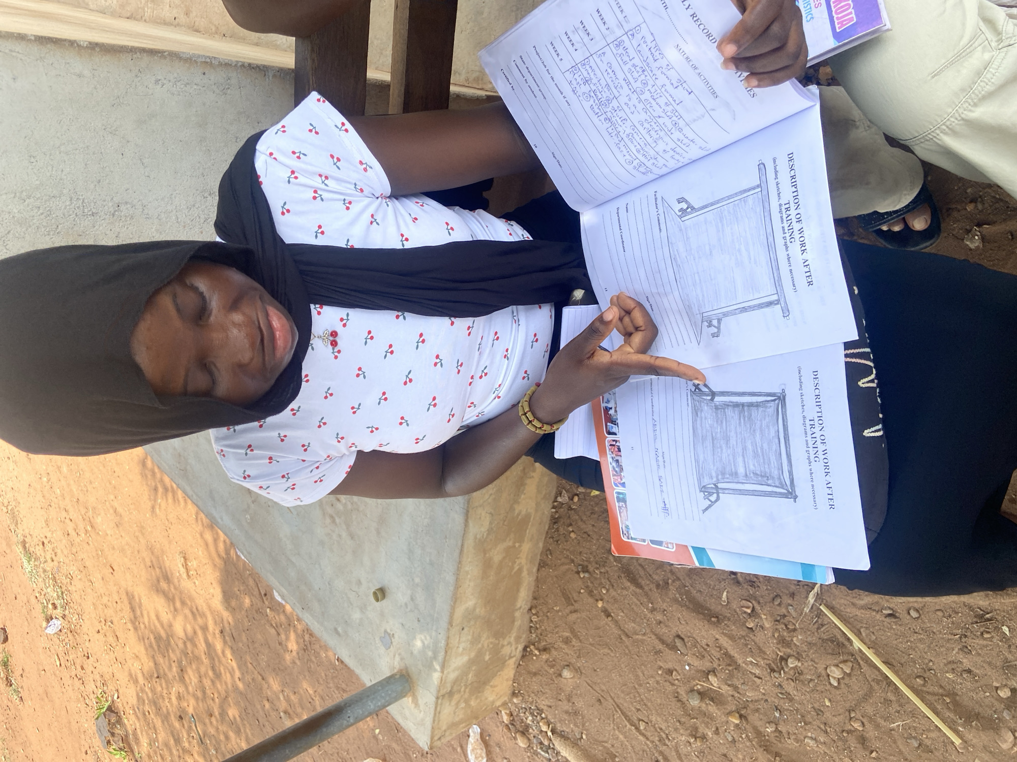 A person is seated outside on a cement block in Lokoja, Nigeria, holding two open notebooks or workbooks. The person is wearing a white shirt with red cherries, a black headscarf, and a black scarf around their neck. They are pointing to a diagram of a tent or shelter drawn in one of the open workbooks. The other workbook contains handwritten notes under the heading "NATURE OF ACTIVITIES" and lists weeks 1-5 with corresponding actions. The environment is dirt and sparse foliage. Another person is partially visible in the upper right corner.