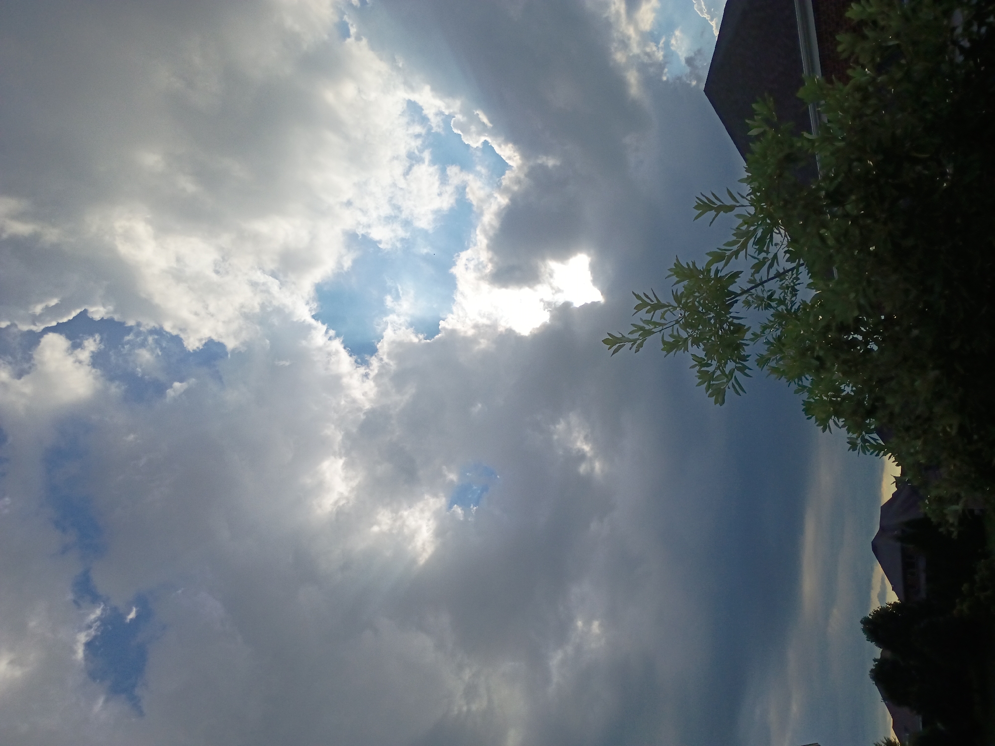 The image captures a cloudy sky with shafts of sunlight breaking through. The foreground features the dark silhouettes of trees and the roofs of houses. The lower portion of the image shows a residential area with houses partially obscured by foliage. The sky is dynamic, with dark storm clouds mixed with brighter, illuminated patches where the sun is visible. The scene appears to be from a suburban environment.