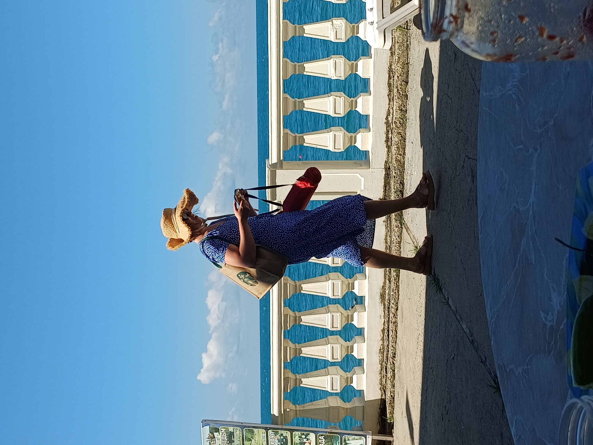 A woman wearing a blue polka dot dress and a wide-brimmed straw hat walks along a paved promenade by the sea in Sochi, Russia. She is carrying a large tote bag on her left shoulder and a rolled-up red mat in her right hand. The promenade overlooks the Black Sea, with a white balustrade separating it from the water. In the foreground, a table is partially visible, covered with a blue and white patterned cloth. On the table, there is a glass jar with some red contents. A white beach chair is visible in the far right of the image. The sky is a clear, bright blue with a few scattered white clouds.