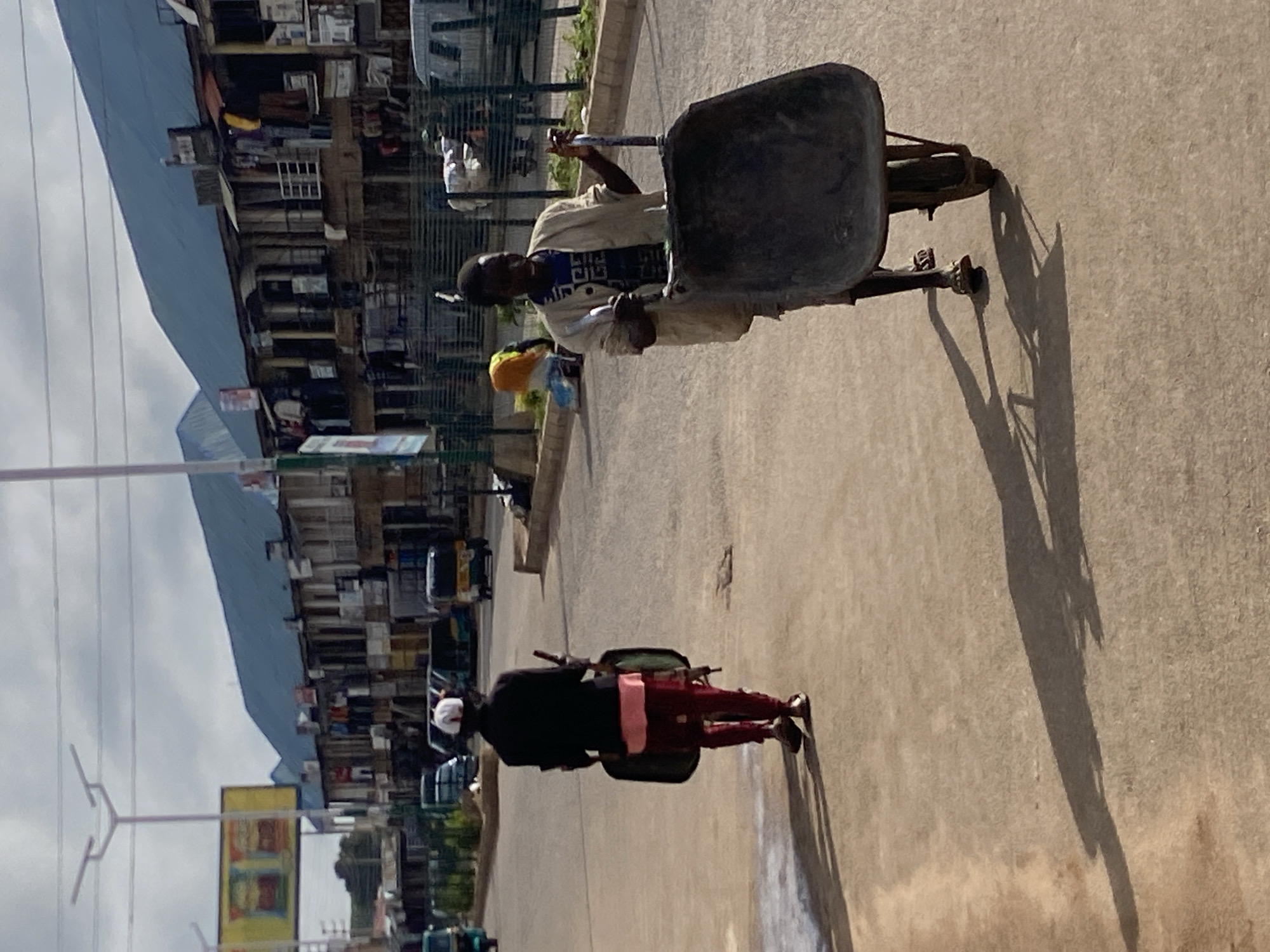 In the city of Bukuru, Nigeria, two individuals are shown pushing wheelbarrows on a paved street lined with multi-story buildings. The building on the right is a commercial structure with shops and balconies. The man in the foreground, wearing a light-colored jacket over a blue t-shirt and shorts, is actively pushing a large, dark-colored wheelbarrow. He is looking forward, with his hands on the wheelbarrow's handles. The second individual, further down the street and facing away from the camera, is dressed in a dark jacket, pink shirt, and dark trousers, also pushing a wheelbarrow. The street is well-lit by natural sunlight, casting long shadows. Several vehicles, including a tricycle and what appears to be a van, are visible in the background, suggesting an active urban environment. The sky is overcast with clouds.