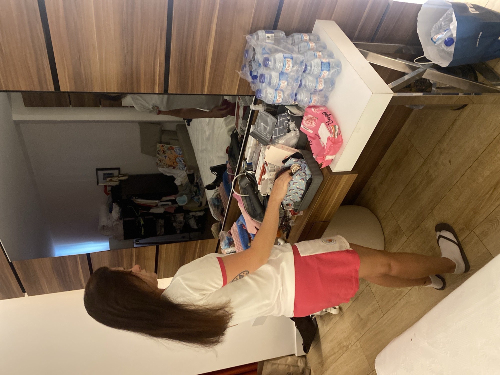 A young woman with long dark hair, wearing a white t-shirt with pink trim and a pink skirt, stands facing away from the camera in a bedroom. She is looking at a vanity with a large mirror. On the vanity, there are several bottles of water, a pink package of wet wipes, and various items in containers. The room has wooden accents and a wooden floor. To the right of the vanity, there is a blue paper bag in the corner. The reflection in the mirror shows a closet filled with clothes and a bed with bedding. The scene appears to be in Aksu, Turkey.