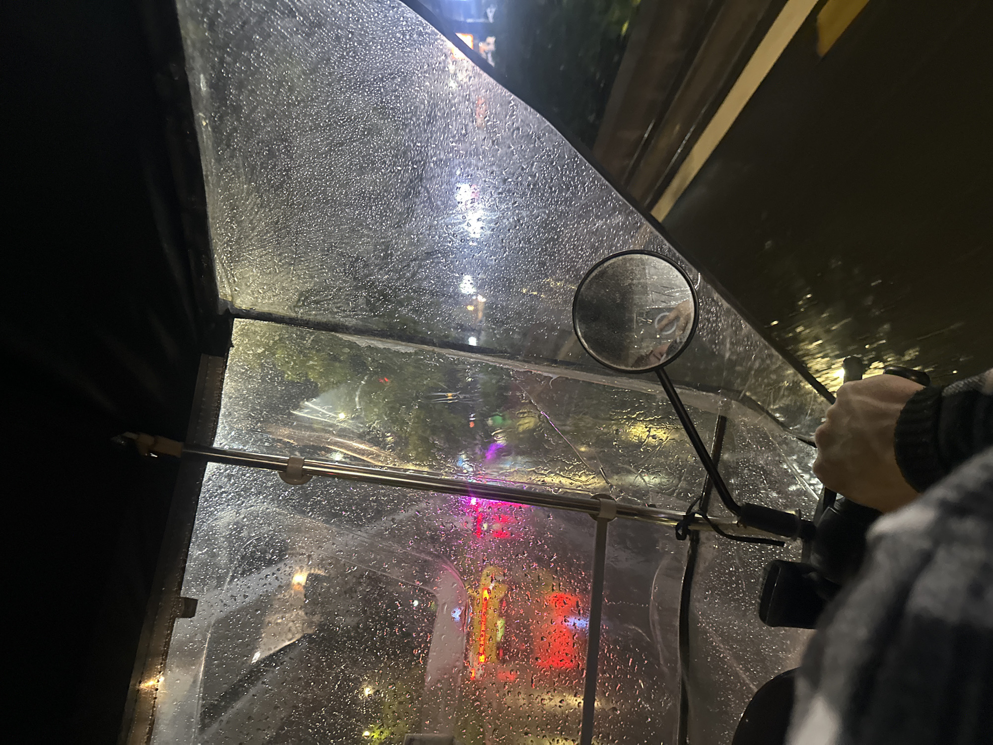 The image is taken from inside a vehicle, likely an auto rickshaw, on a rainy night. The windshield is covered in raindrops, blurring the view of the Chongqing High-tech Zone. Outside, various lights are visible, including streetlights and illuminated signs in red and yellow hues. A side-view mirror is present on the right side of the windshield. A hand wearing a patterned sleeve is visible, presumably holding the vehicle's controls. The overall environment suggests a nighttime setting with active city lights and rainy weather conditions.