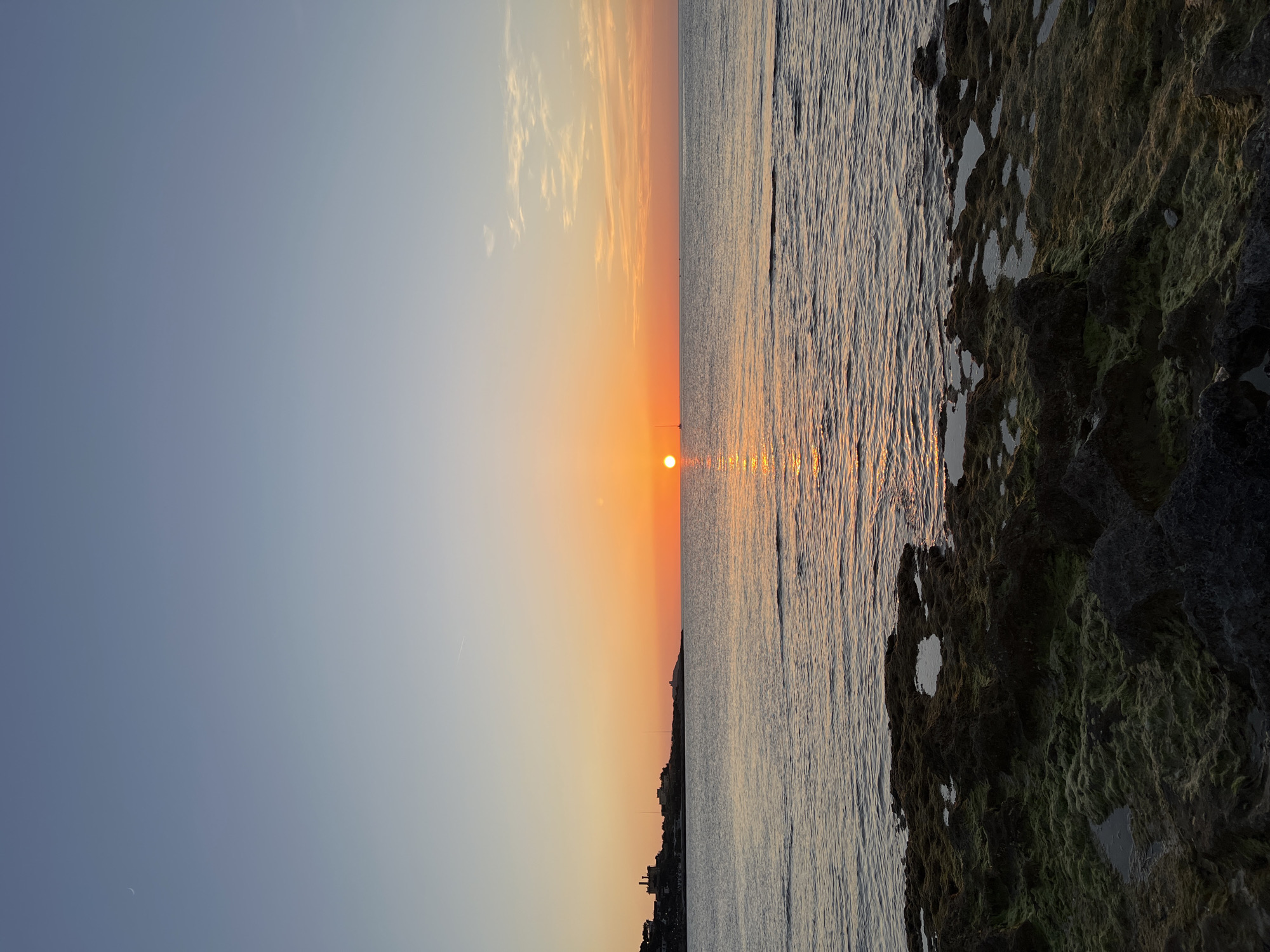 A serene sunset over the Mediterranean Sea near Palermo, Italy, is captured in this image. The sun, a vibrant orange orb, is low on the horizon, casting a warm glow across the water and the sky. The sea reflects the sunset's colors, with a shimmering path of light leading towards the viewer. In the foreground, dark, textured rocks covered in green algae and small pools of water form the rocky coastline. To the left, a distant silhouette of buildings on a hillside suggests a coastal town. The sky transitions from a pale blue at the top to warm orange and yellow hues near the horizon, with a few wispy clouds scattered across the expanse. A faint crescent moon is visible in the upper left corner of the sky. No people are present in the scene.