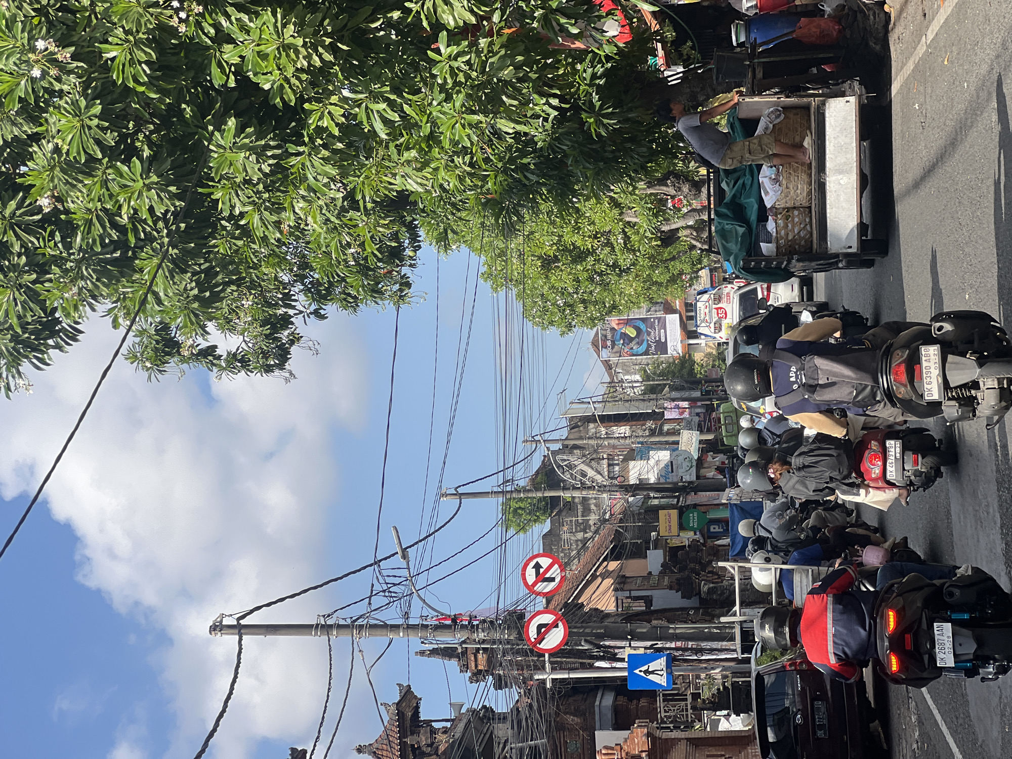 The image shows a street scene in Denpasar, Indonesia, with traffic congestion and a vibrant urban environment. Several motorcycles are visible, some carrying multiple passengers. A truck loaded with baskets is also present, with a man standing on its side. The street is lined with buildings, some with traditional Balinese architecture, and decorated with intricate carvings. Overhead, a complex network of electrical wires crisscrosses the sky, typical of many Asian cities. A blue pedestrian crossing sign is mounted on a pole, along with traffic signs indicating no left and no right turns. Lush green trees with white flowers line one side of the street, providing shade and a touch of nature amidst the urban hustle. The sky is a clear blue with scattered white clouds, suggesting a sunny day. The overall impression is of a busy, dynamic street in a tropical city.