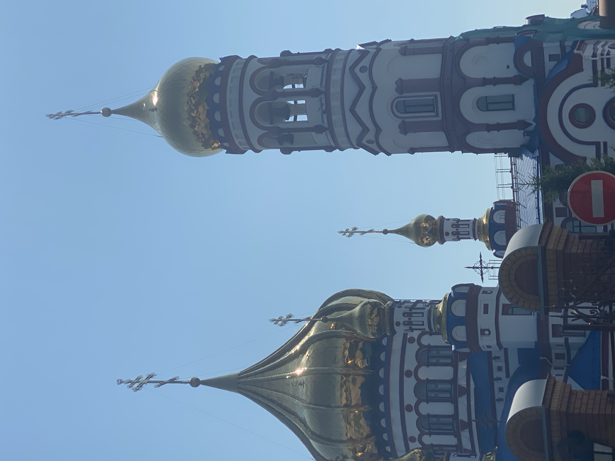 The image shows a view of the Cathedral of the Transfiguration in Novokuznetsk, Russia, under a clear blue sky. The cathedral features multiple golden domes topped with Orthodox crosses, and its architecture includes white walls with red trim and blue accents. A prominent bell tower with arched openings is visible on the right side of the frame. In the foreground, parts of a fence and a red and white circular sign are visible. The image captures a sunny day with bright sunlight reflecting off the golden domes.