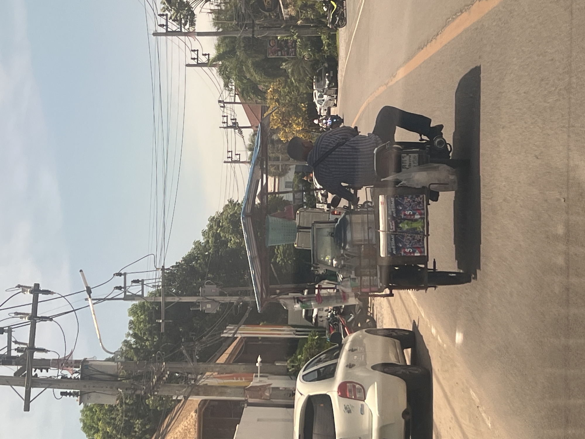 In the city of Ko Samui District, Thailand, a man in a blue and white striped shirt and dark pants is riding a motor-tricycle down a paved road. The motor-tricycle is loaded with items, including what appears to be a blue plastic basket and several bottles. The man is positioned in the driver's seat, facing away from the camera. The road is marked with a yellow line on the right side. In the background, there is a white car parked on the left side of the road. The environment features a clear blue sky, overhead power lines supported by wooden poles, and lush green trees. The scene appears to be a street in a tropical urban area.