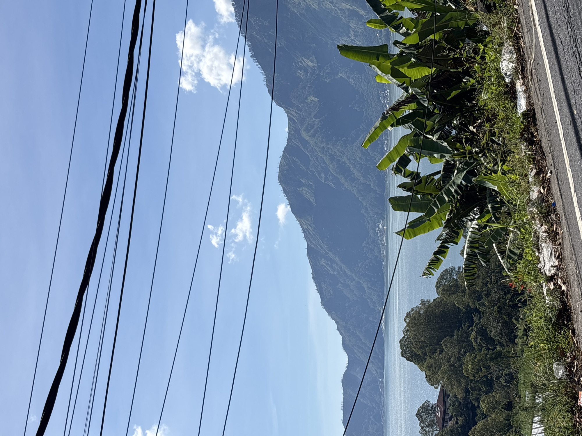 The image depicts a landscape in Kintamani, Indonesia, featuring a view of Mount Batur across Lake Batur under a blue sky with scattered clouds. Power lines are prominent in the foreground. Lush tropical vegetation, including banana trees and other greenery, grows along the roadside. A segment of the paved road is visible to the right.