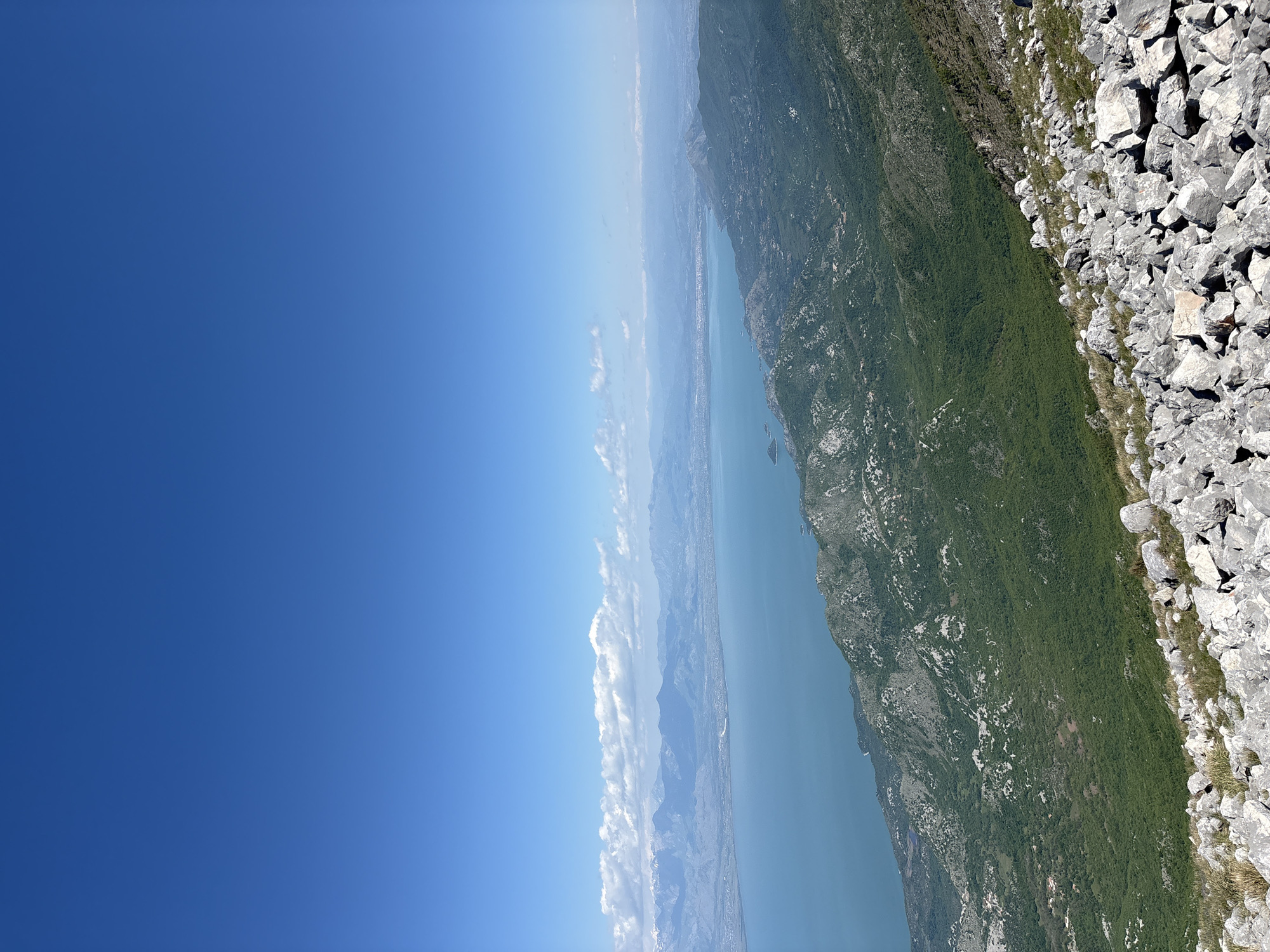 The image shows a view from a rocky mountain edge overlooking a long, narrow body of water, likely a bay or fjord, with green, tree-covered slopes rising on the opposite side. The sky is a clear, bright blue, with a line of clouds along the horizon and at left of the image. The rocky edge in the foreground is composed of large, light-colored rocks. The location is Mali Mikulići, Montenegro.