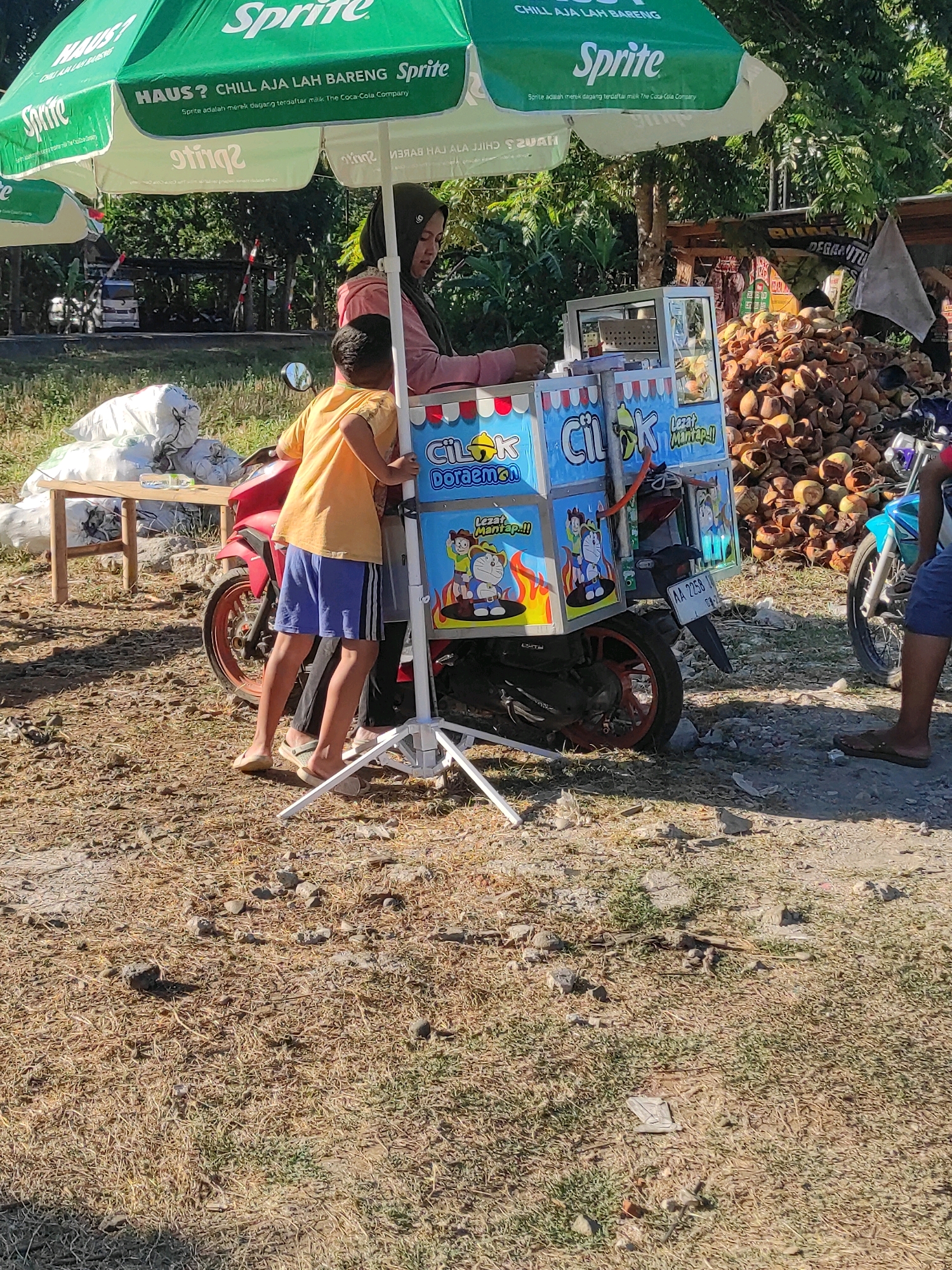 The image depicts a street scene in Purwodadi, Indonesia, with a food cart attached to a red motorcycle. A woman in a hijab and a young boy are attending to the cart, which is decorated with cartoon characters, including Doraemon. The cart appears to be selling snacks or drinks. The motorcycle is parked on a dirt road, with a pile of coconuts in the background. A large green umbrella, branded with "Sprite," provides shade over the food cart. The overall environment suggests a sunny day in a rural or semi-urban area.