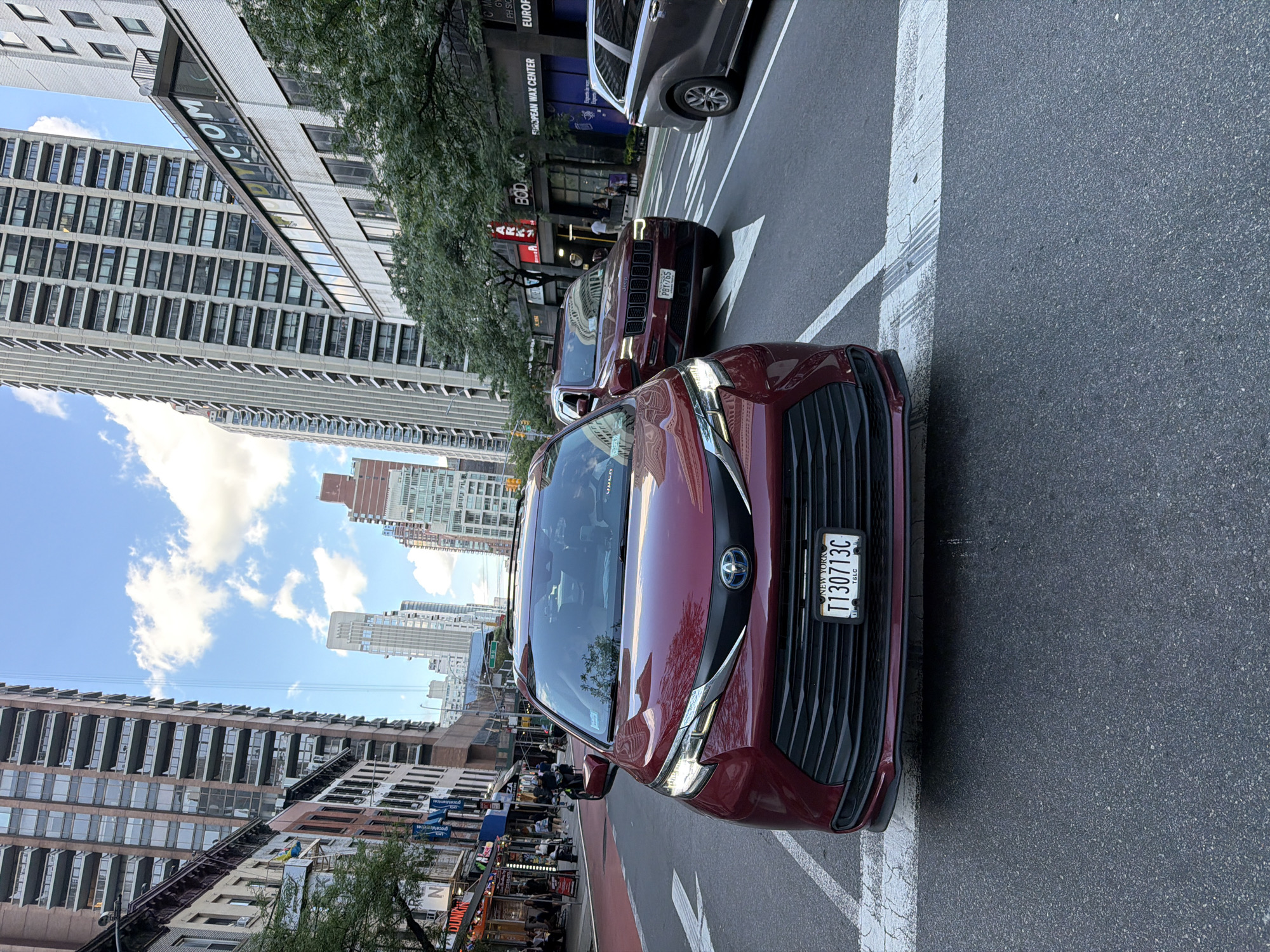 In New York City, USA, a red Toyota Sienna is stopped at a crosswalk on a city street, facing the camera. The minivan has its headlights on and a New York license plate that reads "T130713C". To its right, a dark red Jeep Grand Cherokee is also stopped, its license plate reading "PBY-765". Behind the Jeep, a gray SUV is visible. The street is lined with tall buildings, including apartment buildings and commercial establishments such as "Dunkin'" and a "European Wax Center". The sky is partly cloudy. Pedestrians and scooters are seen on the sidewalk in the background. The image appears to be taken from another vehicle in traffic.