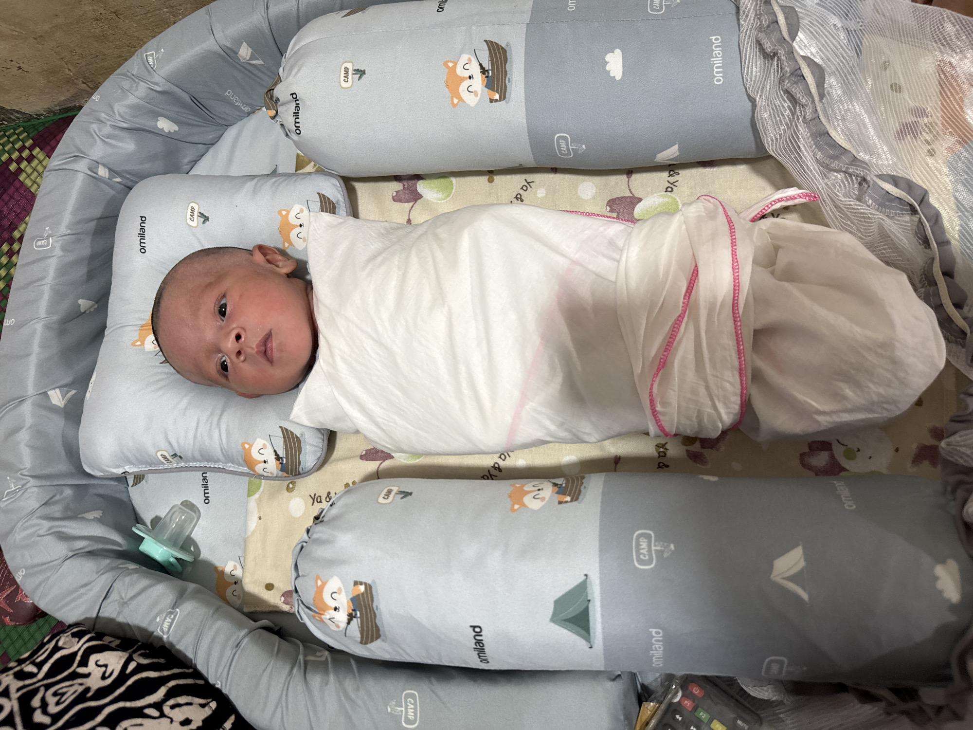 A newborn infant is swaddled in white cloth with pink trim, lying on its back in a crib with grey and blue bumper pads. The bumper pads have a pattern of clouds, tents, and foxes. A pacifier rests on the bumper pad to the infant's right. The background appears to be a crib mattress with a light-colored, patterned sheet. The scene is indoors, likely in a bedroom in Meunasah Raya, Indonesia, given the context.
