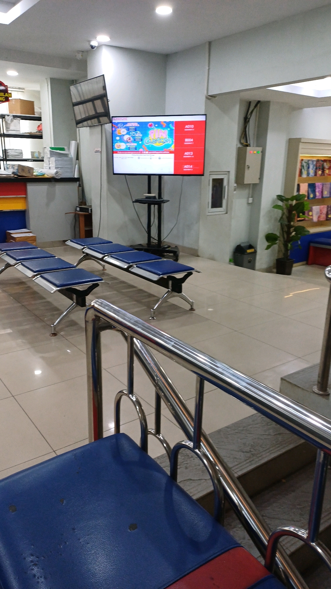 The image depicts the waiting area of a facility, likely a service center or office, in Cibinong, Indonesia. The scene is brightly lit with overhead fluorescent lights. In the foreground, a blue-padded bench with a red accent is partially visible, along with a polished chrome railing leading up a short set of stairs.
Dominating the mid-ground are two rows of identical blue-padded benches, each seating three people. They are arranged parallel to each other, facing away from the camera. The benches have silver metal frames and legs.
A large flat-screen television mounted on a black stand is positioned to the right of the benches, displaying a colorful graphic with text. Above this television, another smaller television or monitor is mounted on the wall, its screen reflecting the room's lighting.
The walls are painted a light grey, and the floor is tiled with large, light-colored squares. To the right of the television, a tall potted plant in a black pot adds a touch of greenery. Further to the right, a beige electrical junction box is visible on the wall, with some indicator lights illuminated. In the background, shelves are stocked with various items, and a blue and red cabinet is visible on the left. The overall impression is one of a clean, functional, and modern waiting space.