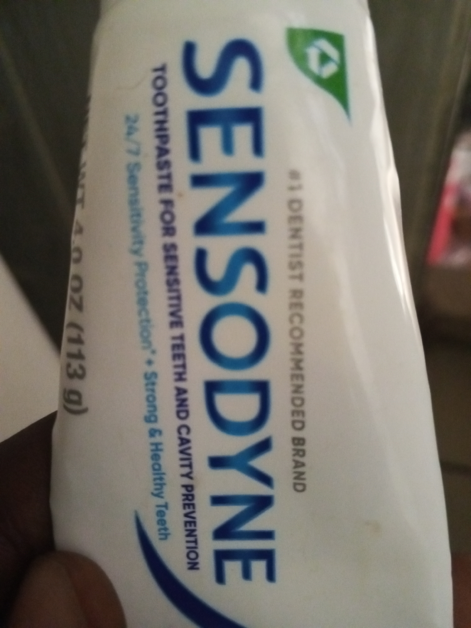 This is a close-up, vertical, eye-level, medium shot of a tube of Sensodyne toothpaste. The tube is white with blue lettering. The brand name "SENSODYNE" is prominently displayed in large blue capital letters. To the left of the brand name, there is text that reads "TOOTHPASTE FOR SENSITIVE TEETH AND CAVITY PREVENTION" and "24/7 Sensitivity Protection + Strong & Healthy Teeth." Below that, the weight is indicated as "NT 4.0 OZ (113 g)". To the right of the brand name, in smaller text, it says "#1 DENTIST RECOMMENDED BRAND" and there is a small green triangular logo with a white recycling symbol inside. The bottom left corner of the image shows a portion of a dark-skinned hand holding the tube. The background is out of focus and appears to be a dimly lit indoor environment. There are no identifiable landmarks or contextual clues to suggest a specific location within a city.