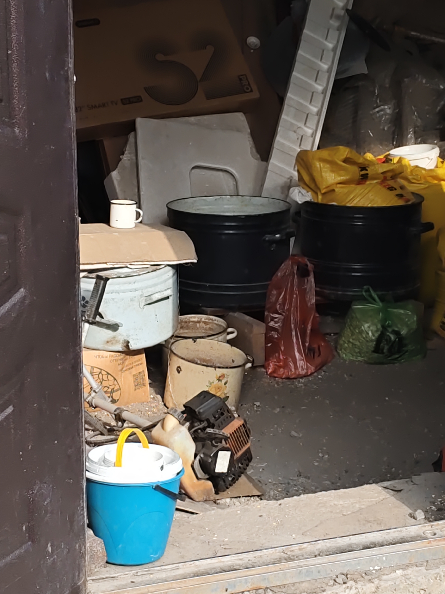 The image shows a cluttered storage area. In the foreground, a blue bucket with a white lid and yellow handle is visible. To its right, a piece of machinery, possibly a small engine, is present. Behind these items, various containers and bags are piled up. There are two large black pots, a light blue metal bin, and several smaller enamel pots with floral patterns. Bags of yellow material and a green bag filled with what appear to be dried leaves or small plants are also scattered around. A red plastic bag lies on the ground between the black pots. A white mug sits atop a cardboard box. In the background, large cardboard boxes are stacked, one with a "32 SMART TV" label. The environment appears to be an indoor space with a concrete floor, likely a shed, garage, or storage room. The location is identified as Nizhnedonskoy, Russia. No people are visible in the image, and no specific actions or interactions are depicted.