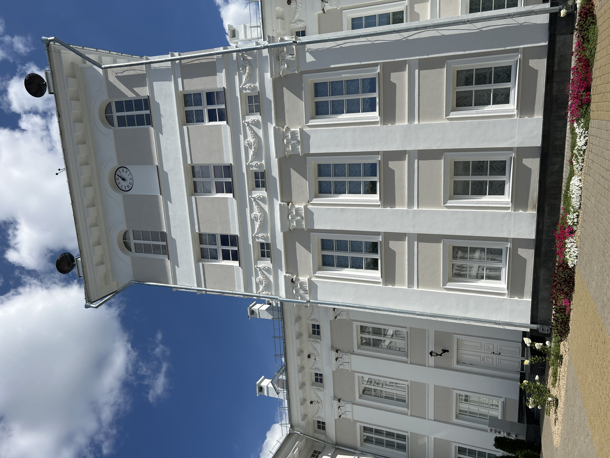 The image shows a multi-story building with a light gray facade and white trim. The building features numerous windows, many of which are arched at the top. A clock face with Roman numerals is visible on the upper portion of the central tower-like section of the building. Decorative architectural elements, such as moldings and columns, are present on the exterior. The building is situated outdoors, with a blue sky and white clouds in the background. In the foreground, there is a paved area and flower beds containing red and white flowers. A drainpipe runs vertically along the side of the building. The scene is devoid of people or any visible activity. The architectural style suggests it is a historical or institutional building. The location is identified as Добасненскі сельскі Савет, Belarus.