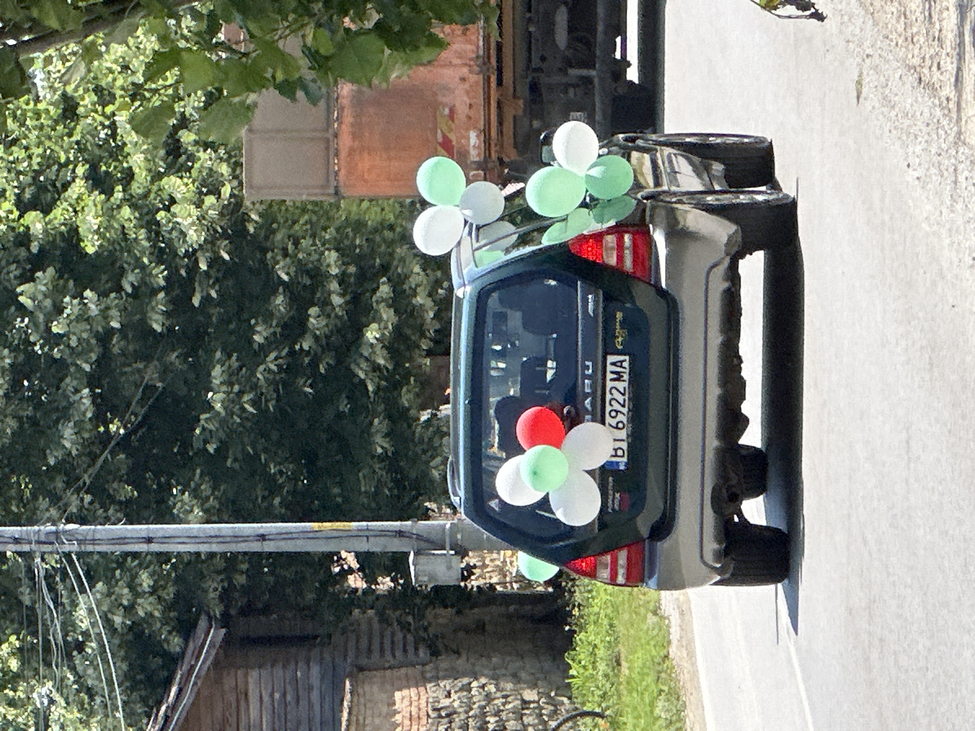 The image depicts the rear of a dark green Subaru Forester with balloons attached to its exterior, suggesting a celebratory occasion. The vehicle is on an asphalt road, with greenery and trees visible in the background, along with buildings and a utility pole. A large truck is partially visible to the right of the Subaru. The license plate of the Subaru reads "BT 6922 MA". The balloons are white, red, and light green, attached to the rear window, trunk, and the side of the vehicle. The scene appears to be in a rural or semi-rural setting, consistent with Prisovo, Bulgaria, a village known for its scenic surroundings and often featuring such decorative elements for events.